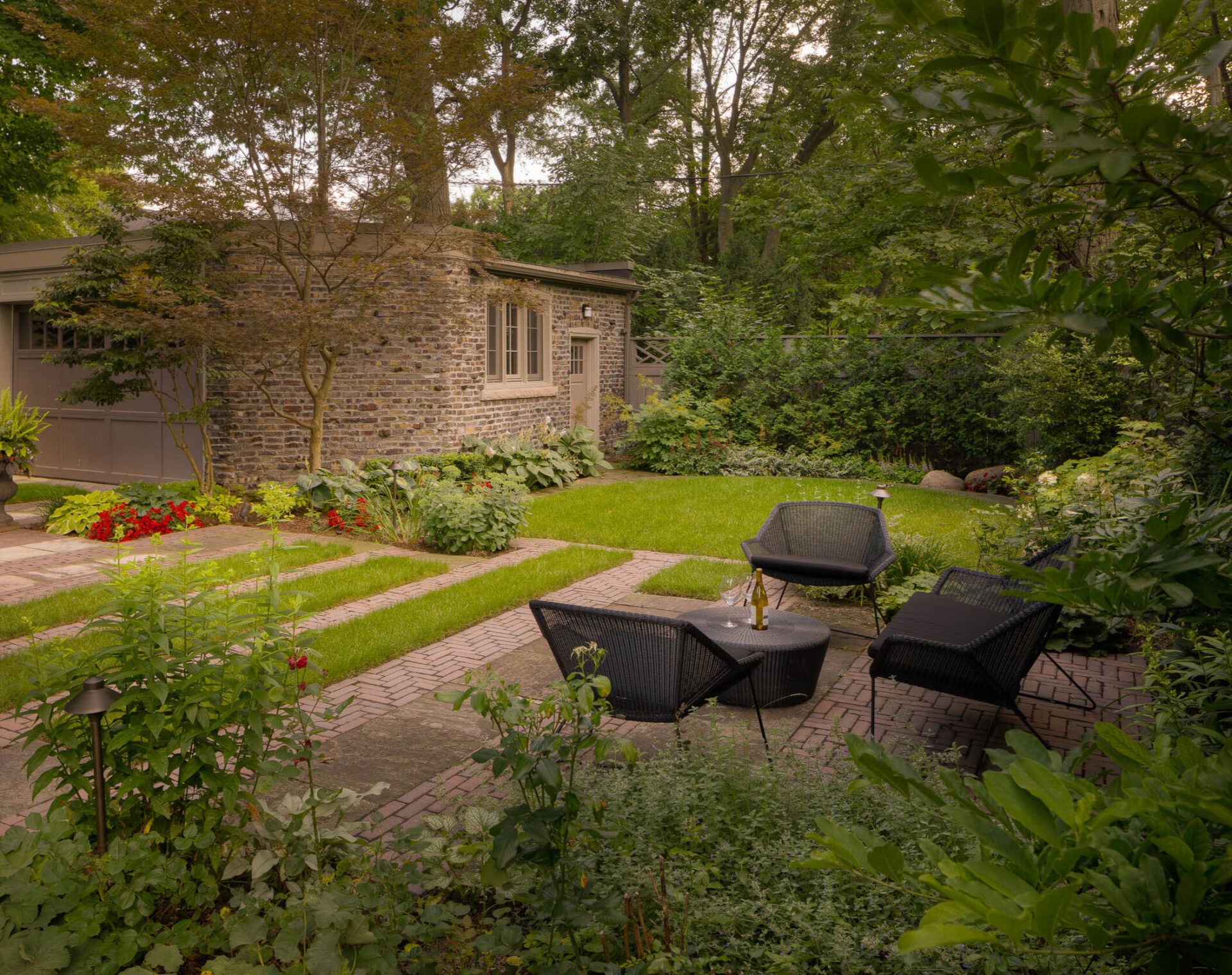 A charming garden with a stone building, surrounded by lush greenery and outdoor seating on a brick patio.