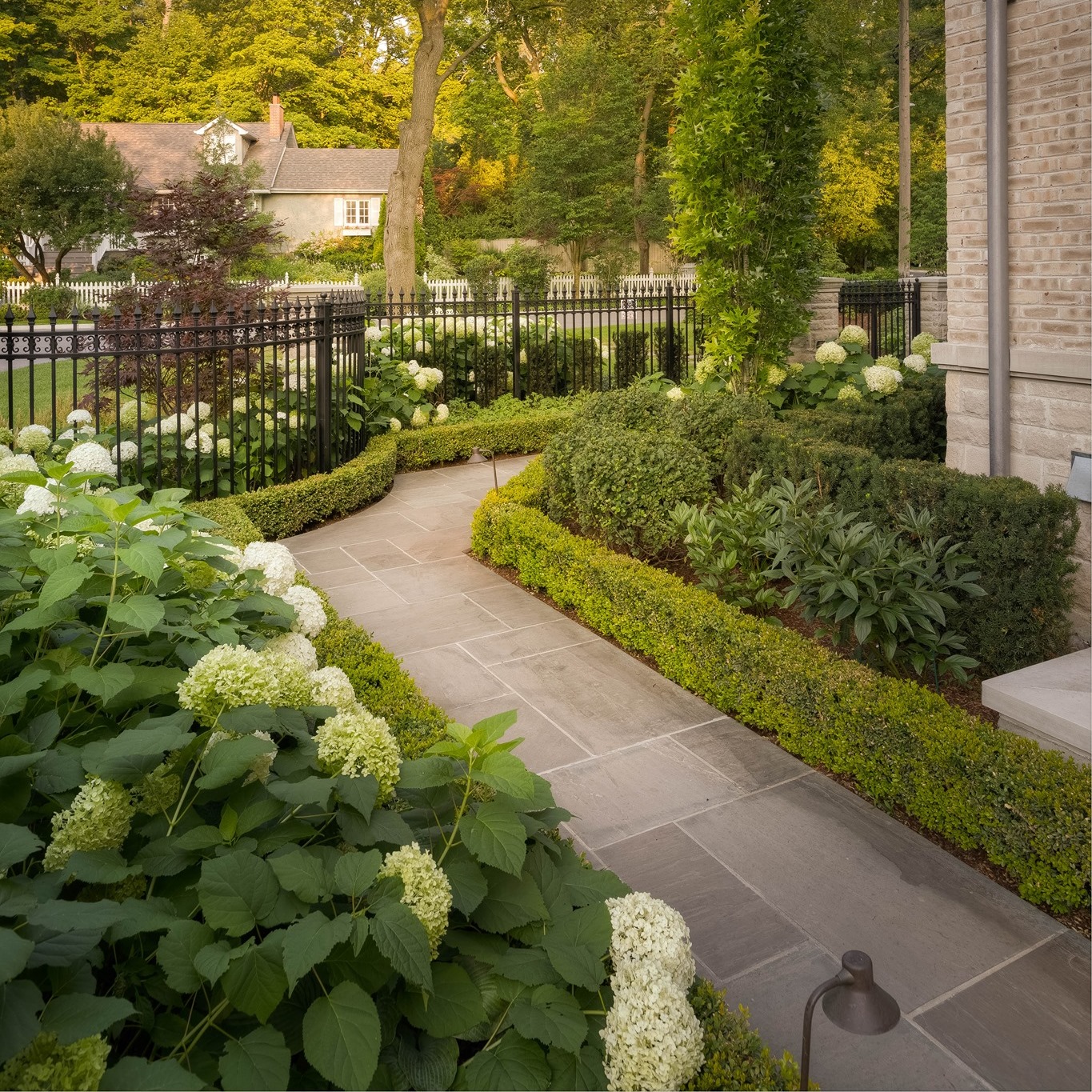 A tranquil garden path with lush greenery and blooming hydrangeas leads to a cozy house, surrounded by trees and a black metal fence.