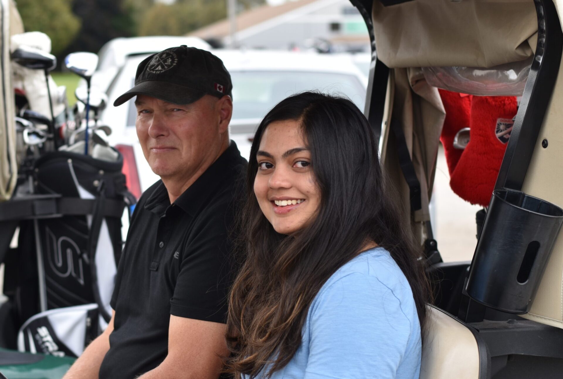 Two people sitting in a golf cart with bags, outdoors. They appear relaxed and content. Trees and cars are visible in background.