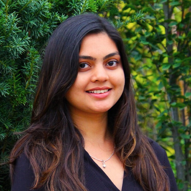 A person with long dark hair smiles outdoors, surrounded by lush green foliage, wearing a black top and a necklace with a pendant.