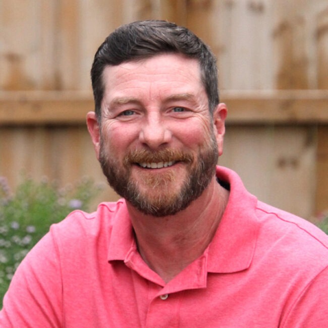 A person wearing a pink shirt smiles at the camera, sitting outdoors with a wooden fence and plants in the background.