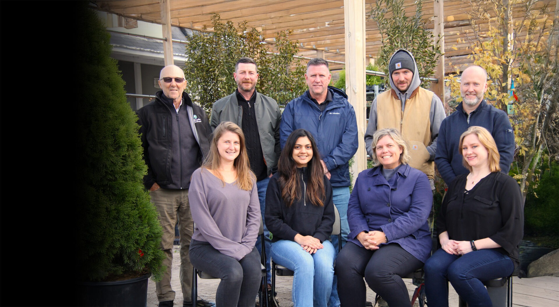 A group of nine people posed outdoors among trees and plants, smiling under a wooden pergola structure with natural surroundings.