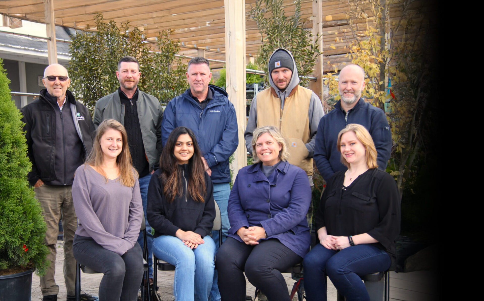 Ten people are sitting and standing in an outdoor setting with greenery and wooden structures, smiling and casually dressed.