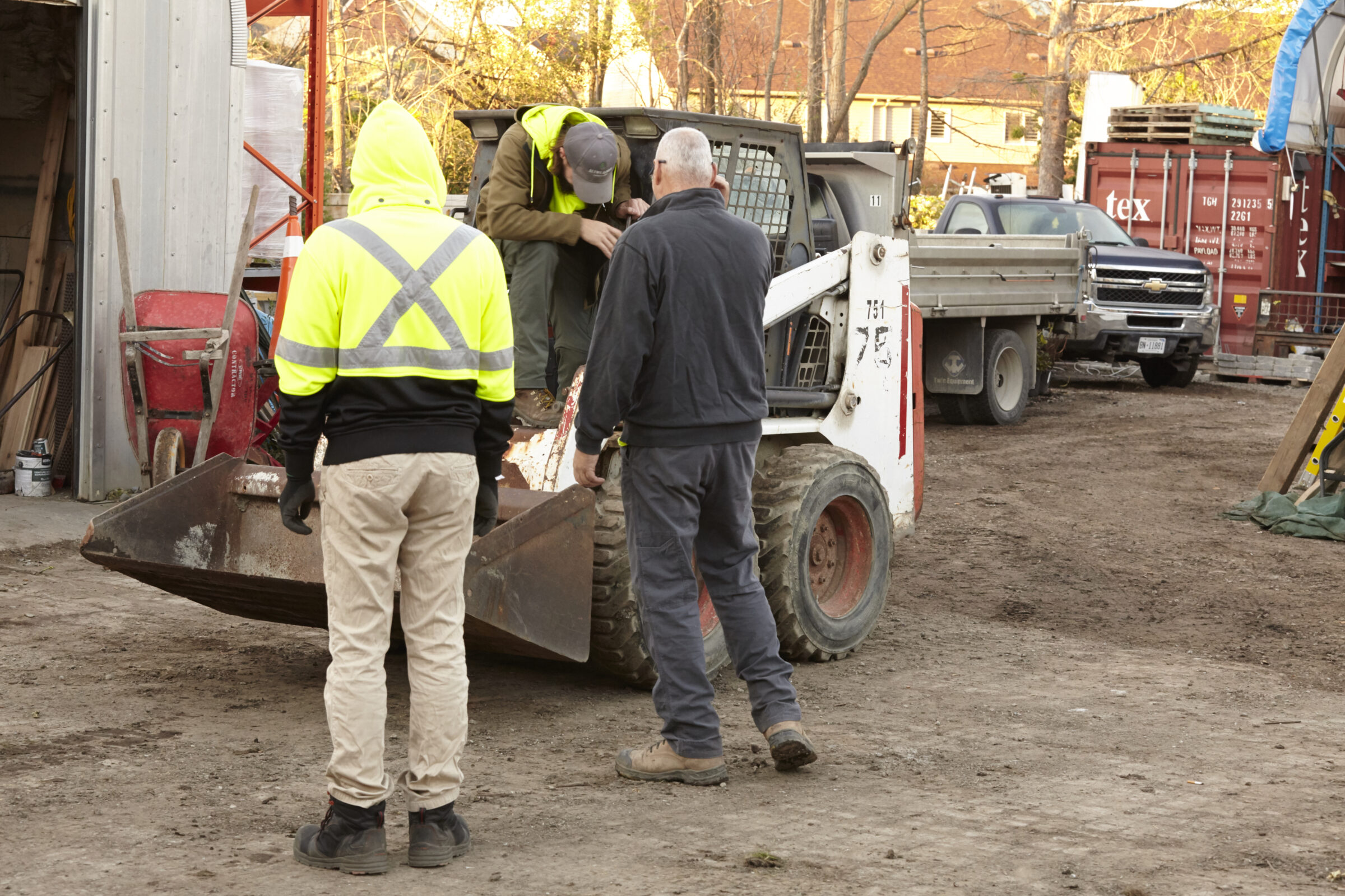 Three people in safety gear work around a small loader at a construction site. Various tools and vehicles are in the background.