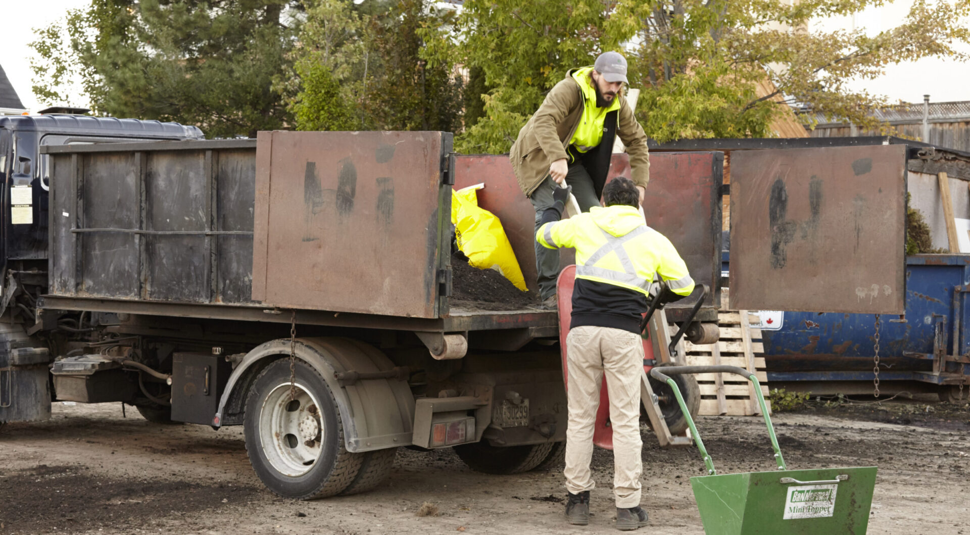 Two people in safety clothing work on loading dirt from a truck into a green wheelbarrow, surrounded by trees and industrial materials.