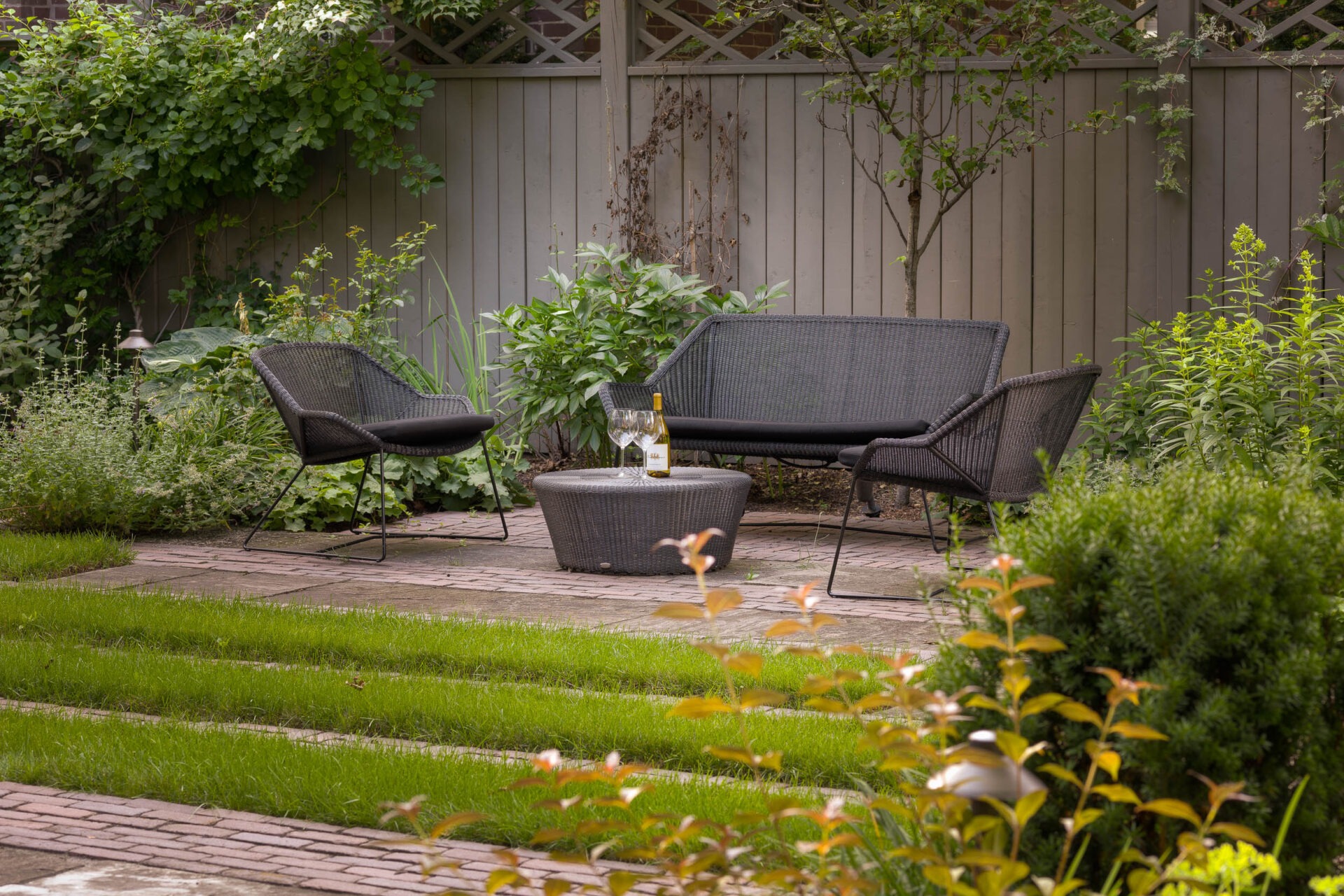 Cozy garden patio with wicker furniture, wine, and glasses on a small table, surrounded by lush greenery and a wooden fence backdrop.