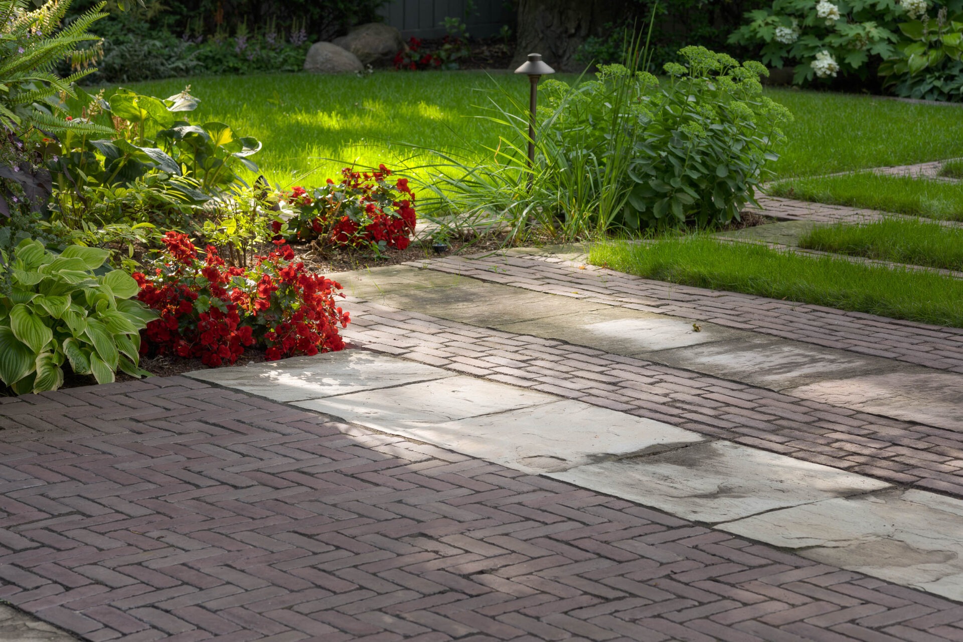 A lush garden with red flowers, green foliage, and a brick pathway lit by sunlight, creating a serene outdoor scene.