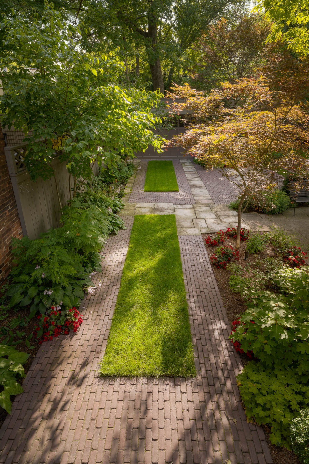 A beautifully landscaped garden path lined with vibrant flowers, lush grass, and overhanging trees. Sunlight creates serene, dappled shadows on the ground.