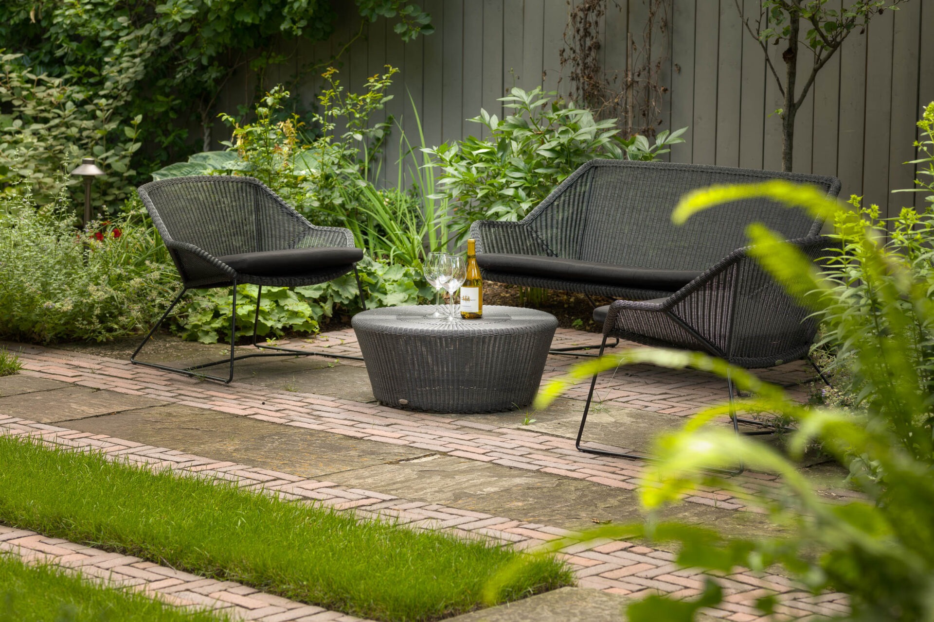 Cozy garden setting with black wicker furniture, small round table, two wine glasses, and bottle. Surrounded by lush greenery and paving stones.