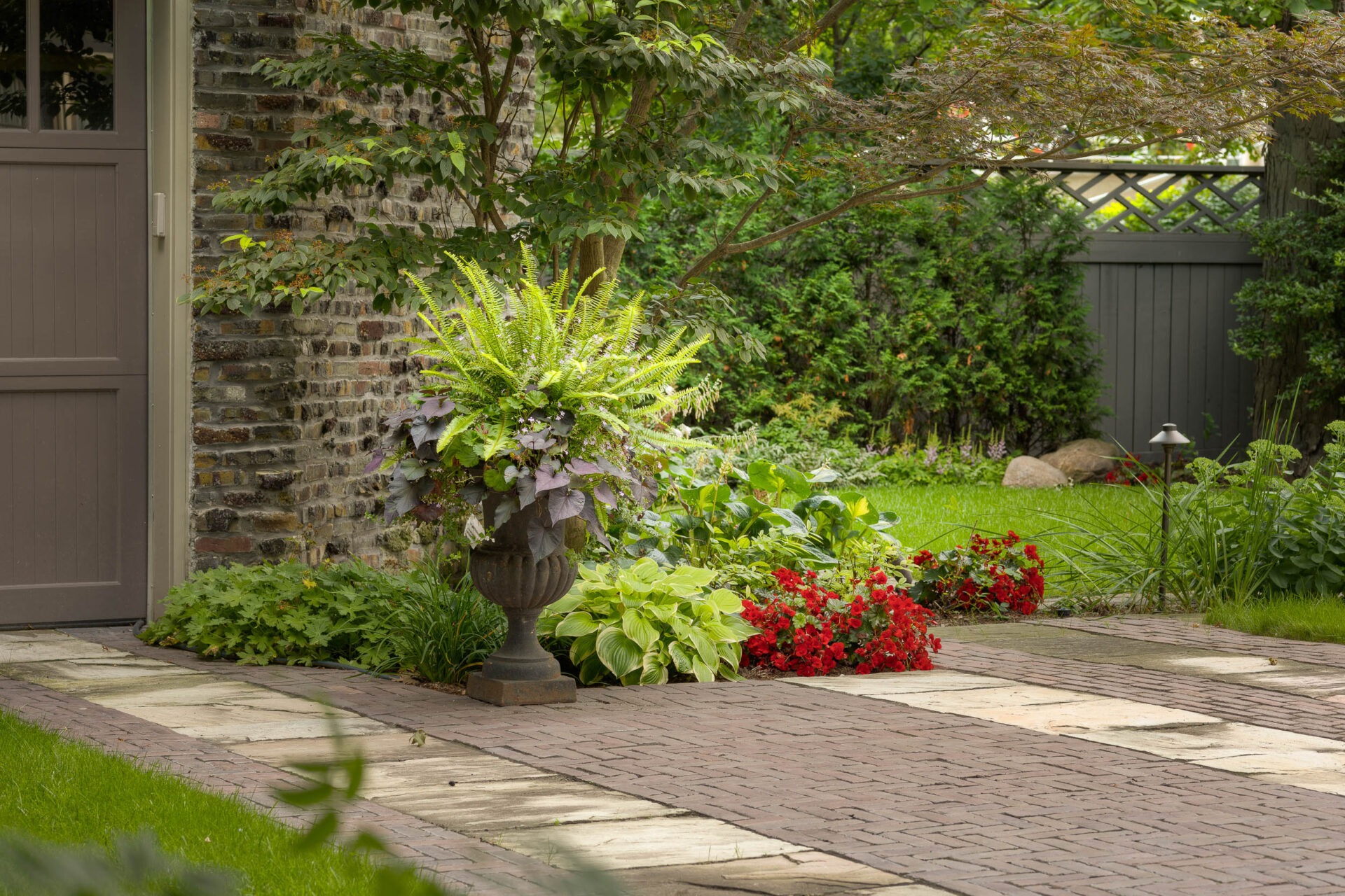 A tranquil garden features vibrant greenery, red flowers, stone pathway, and rustic brick wall, creating a peaceful, natural retreat with lush foliage.