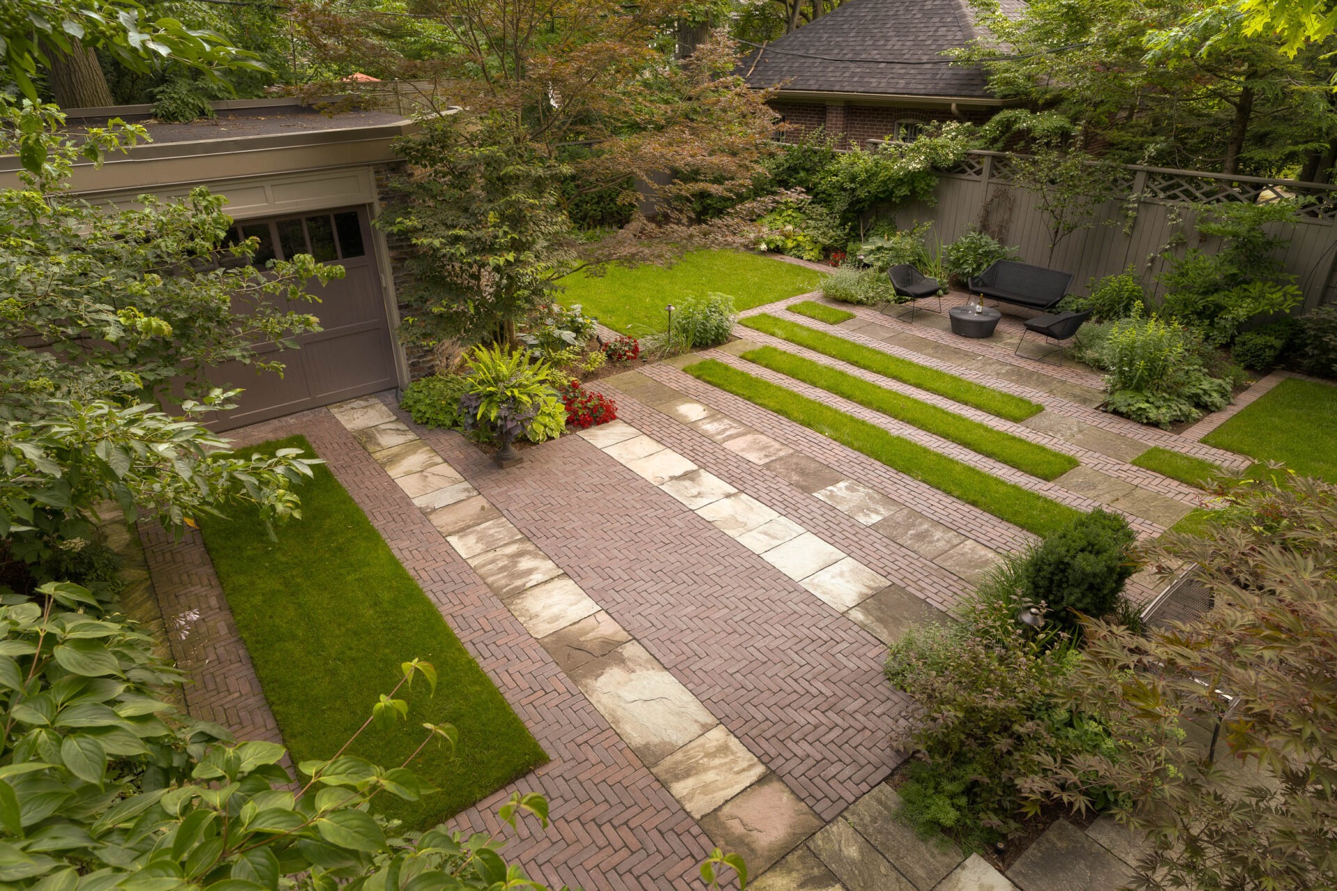 A landscaped backyard with brick and stone paving, lush greenery, and patio seating arranged neatly around a grassy area beside a shed.