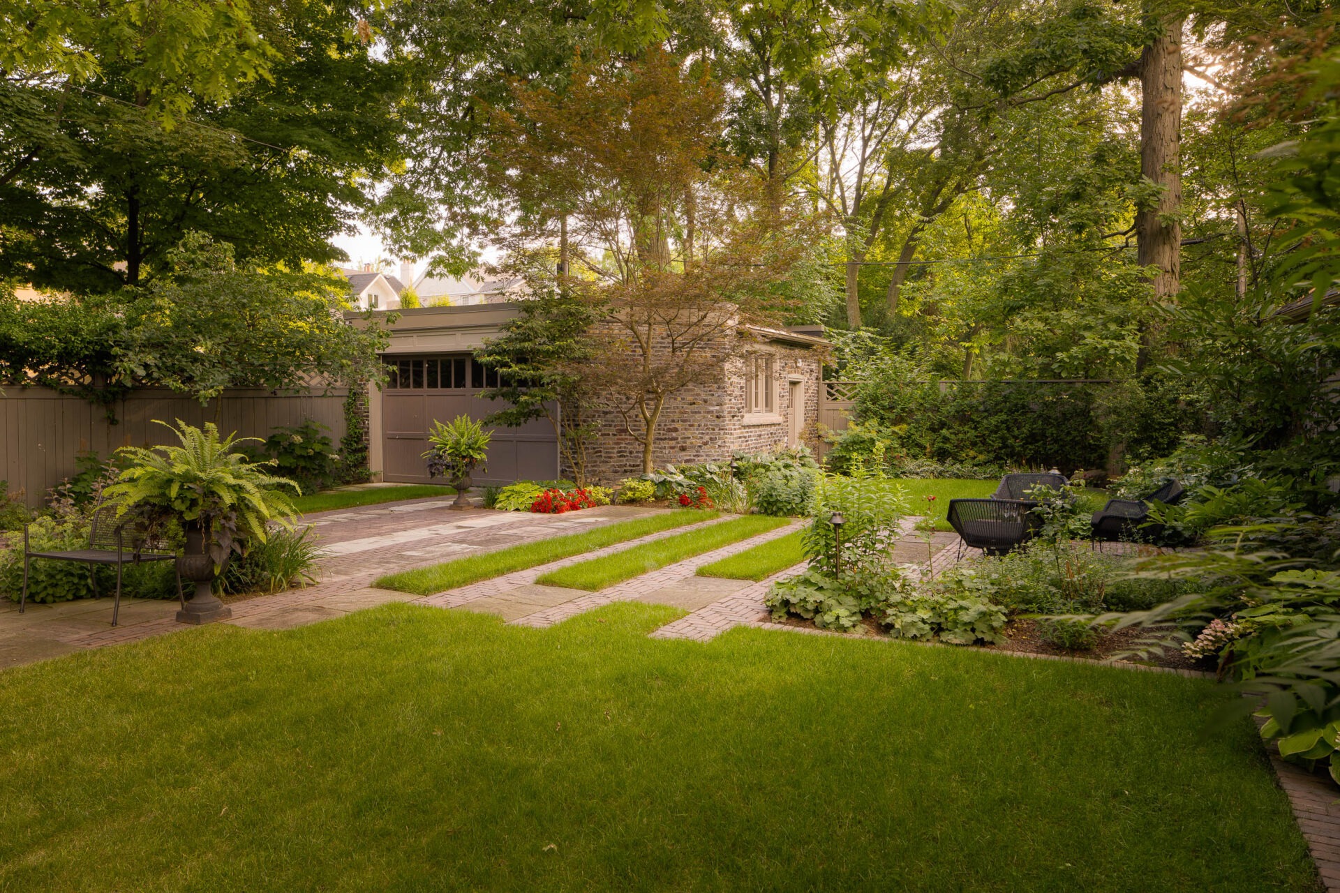 A serene backyard with lush greenery, stone pathways, and a cozy seating area beside a rustic brick building surrounded by trees.