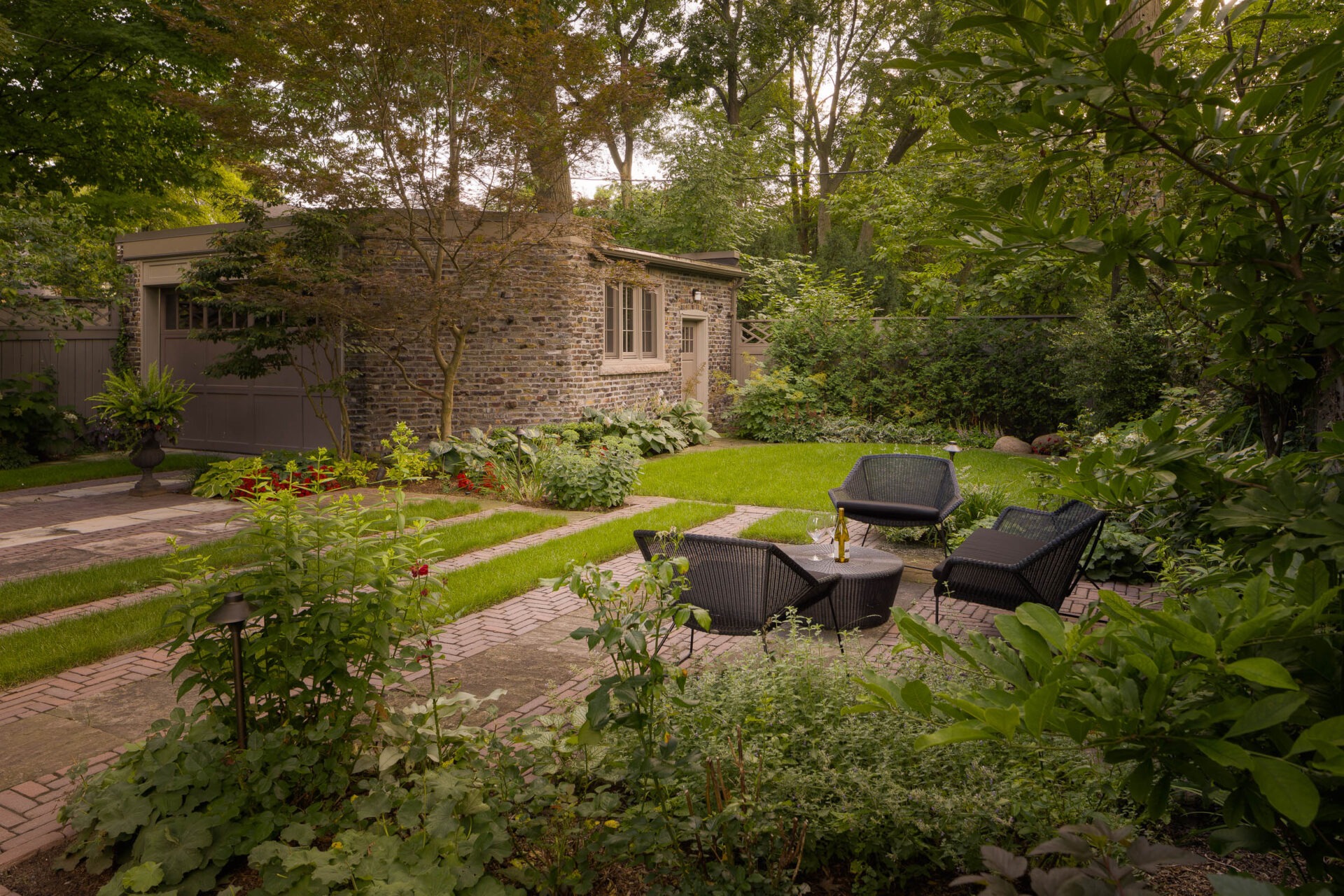 A lush garden features a cozy seating area with black chairs, a brick path, and a quaint brick building surrounded by greenery.