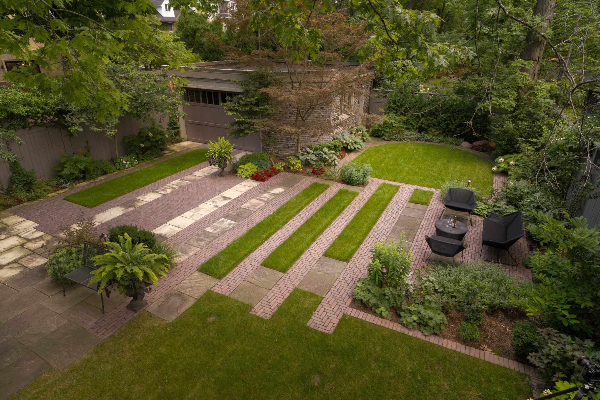 Modern garden with geometric lawn and brick patterns, dense greenery, garden furniture, and a small shed. Peaceful and well-maintained outdoor space.