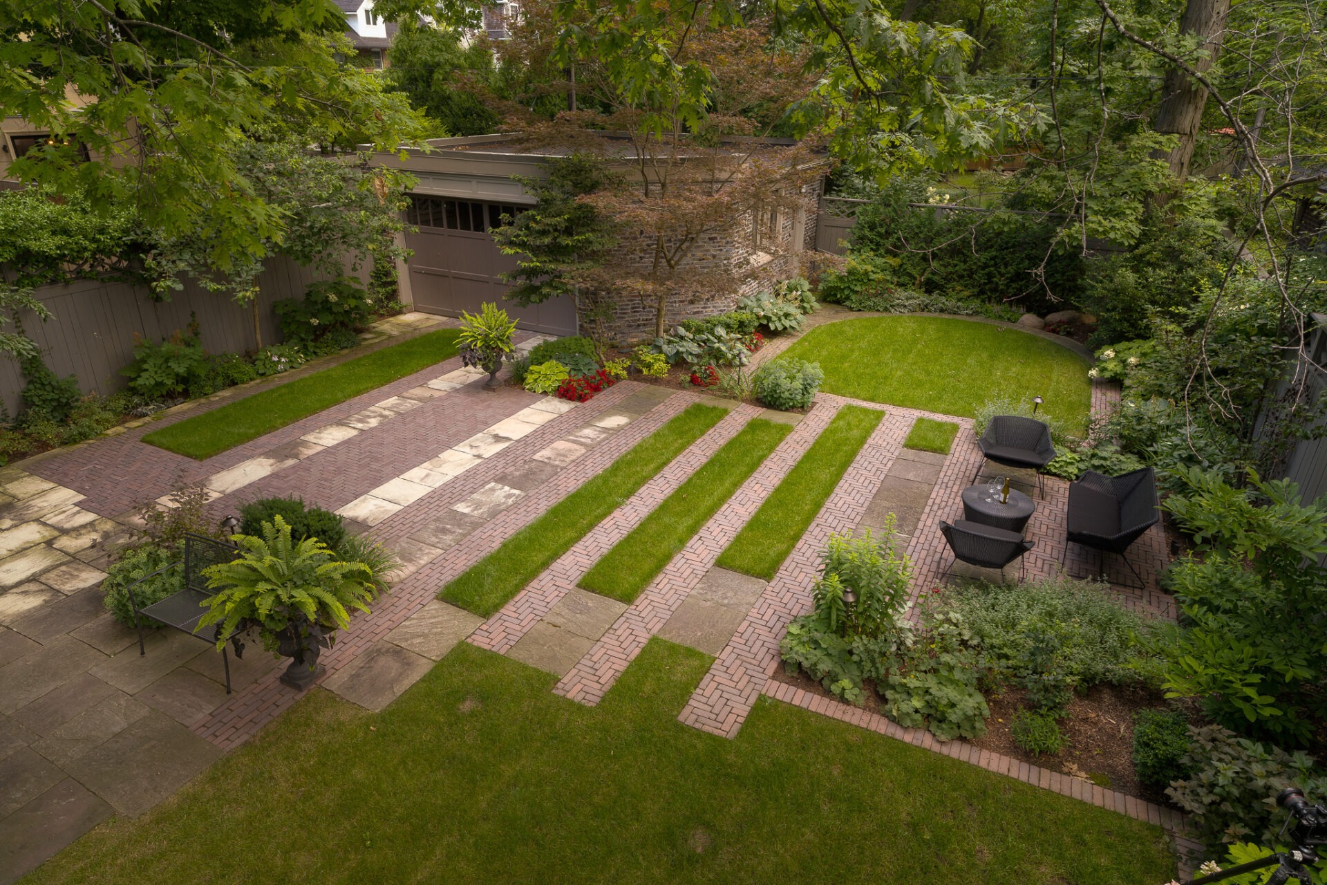 A beautifully landscaped garden with stone and brick pathways, lush greenery, seating area, and a small garage surrounded by trees and plants.