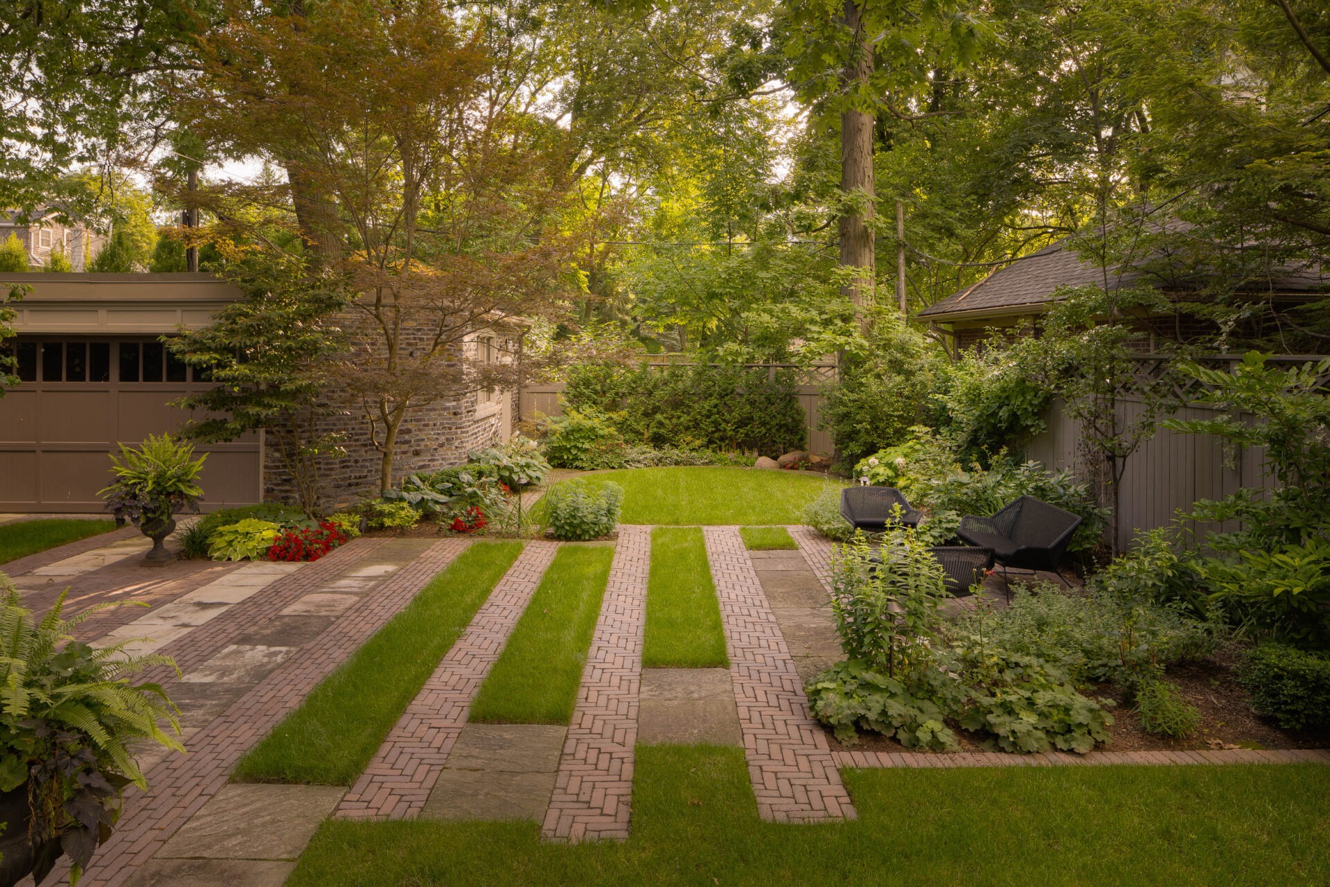 A landscaped backyard features brick pathways, manicured lawn, and lush greenery. Two black chairs and a wooden fence complement the tranquil setting.