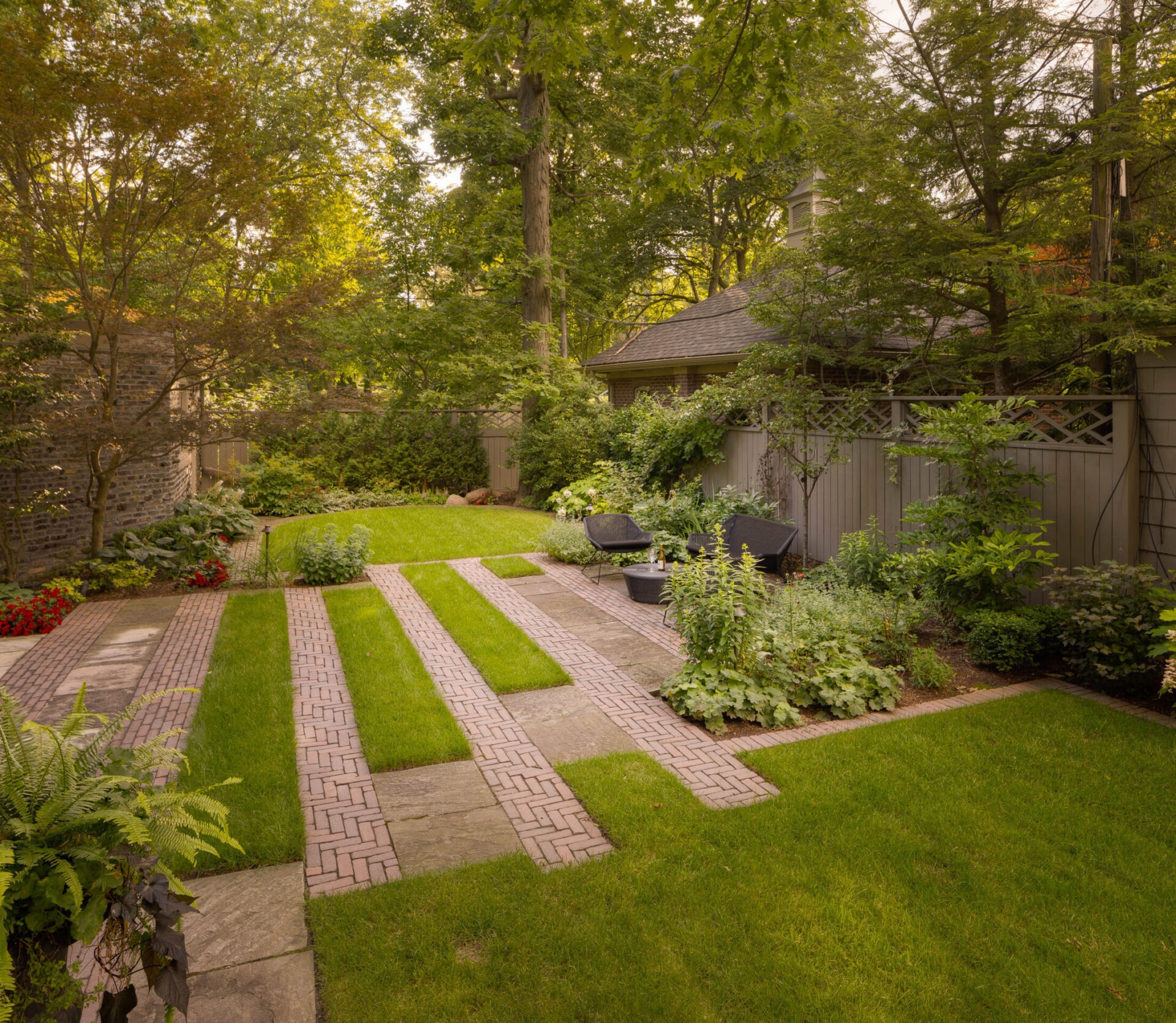 A serene garden features brick pathways, lush green lawns, flowering plants, and patio furniture, surrounded by tall trees and a wooden fence.