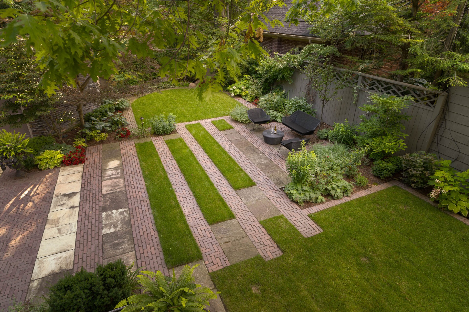 A backyard with brick pathways, lush greenery, and outdoor seating arranged on a patio, surrounded by a wooden fence and trees.