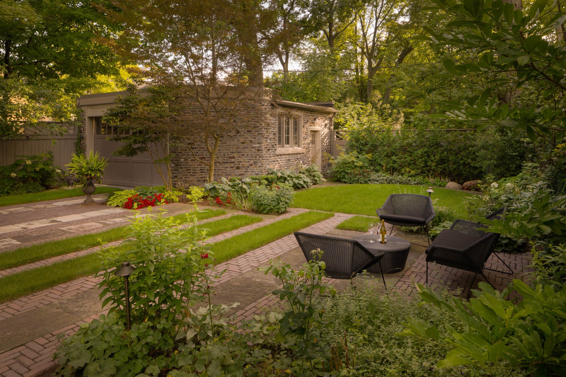 A serene garden with stone pathways, lush greenery, a brick building, and cozy wicker chairs surrounding a table with a wine bottle.