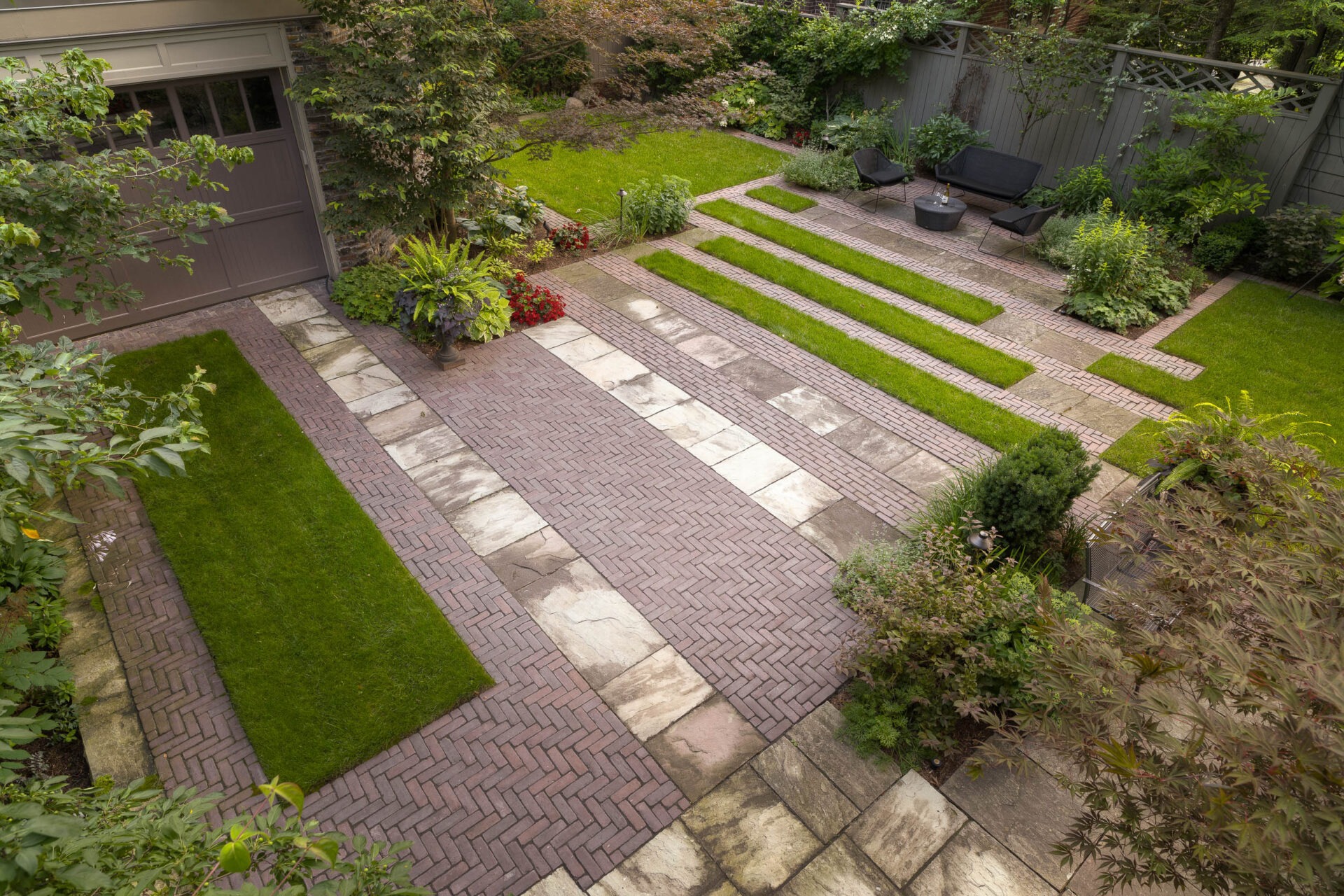 A well-maintained garden features geometric patterns with bricks and grass, surrounded by lush plants and a cozy seating area with chairs.