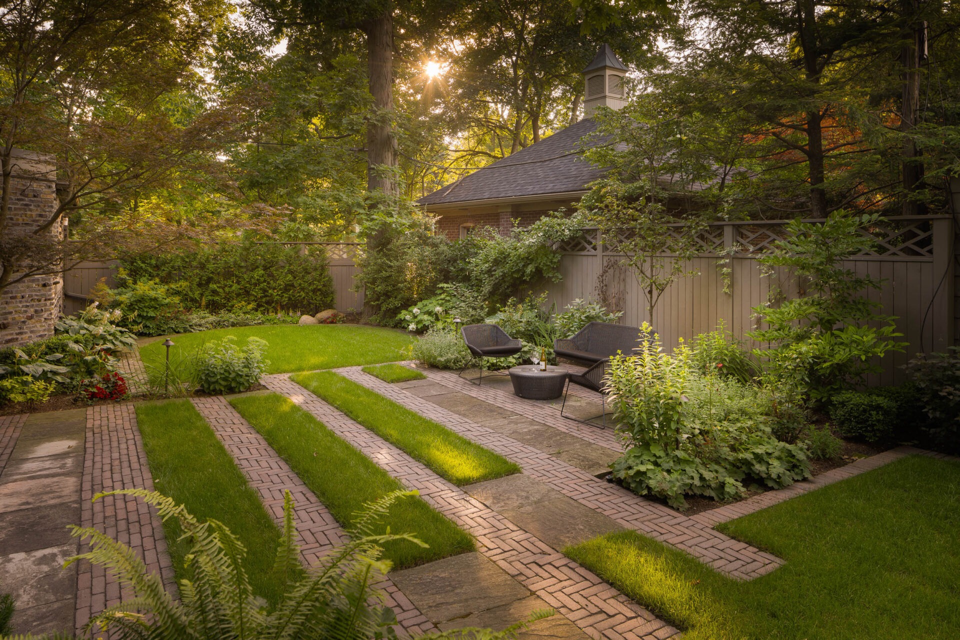 A tranquil garden with brick pathways, lush greenery, outdoor seating, and a small building, illuminated by warm sunlight filtering through trees.