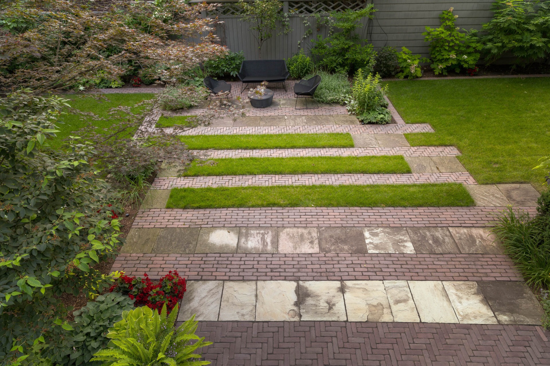 A well-maintained garden with brick pathways, grass strips, patio seating, and surrounding greenery, creating a harmonious outdoor living space.