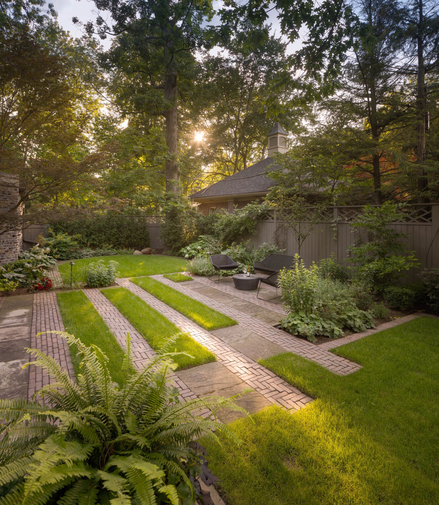 A serene garden features brick pathways, lush greenery, and patio furniture under a canopy of tall trees with sunlight filtering through.