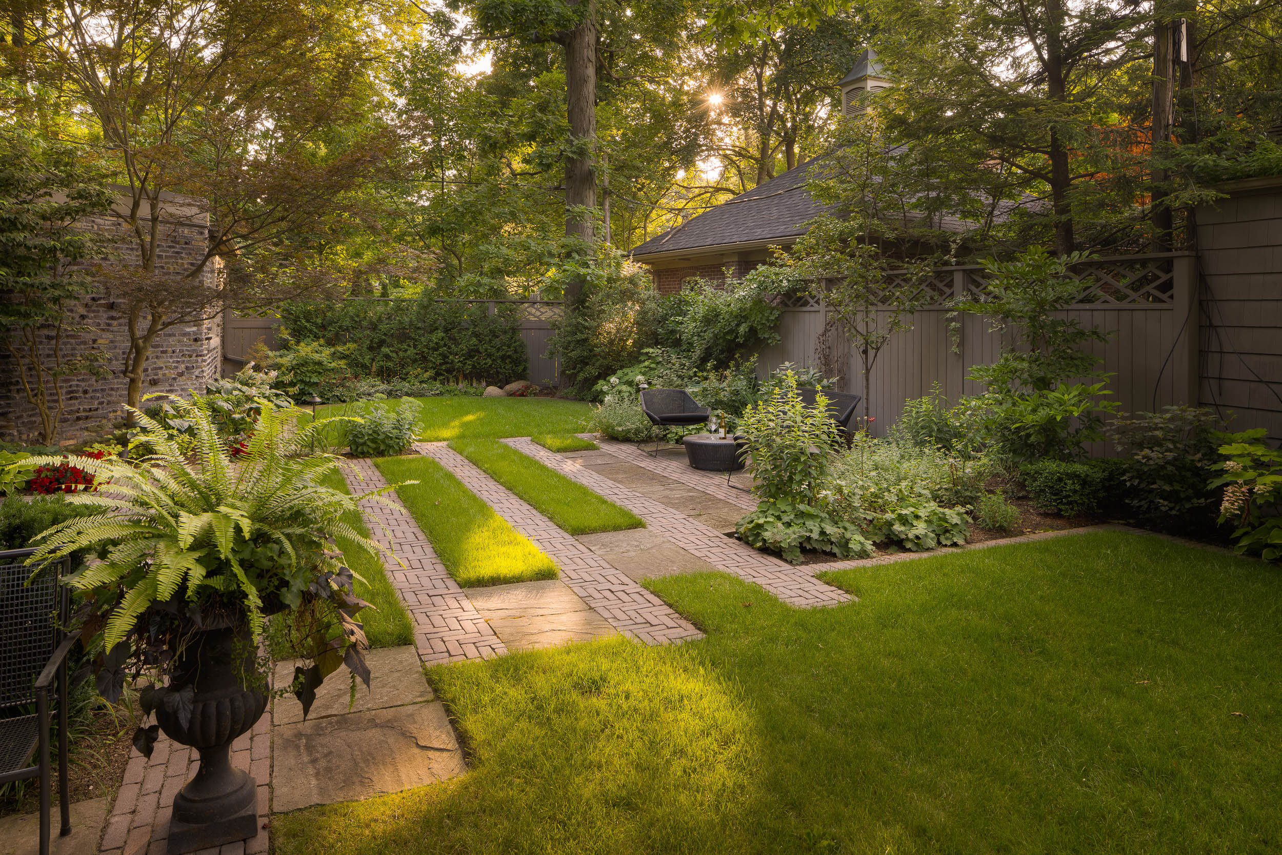 A tranquil garden featuring lush greenery, brick pathways, and patio furniture, surrounded by trees and a rustic stone wall. Sunlight filters through.