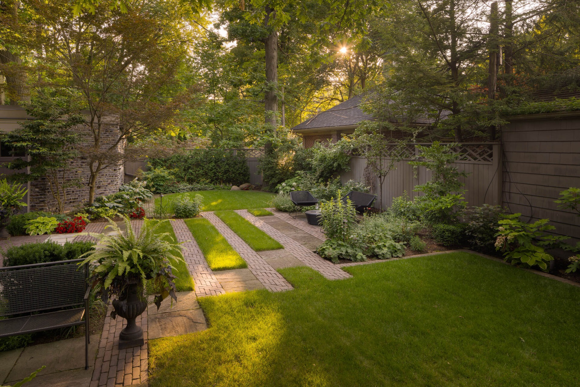 A serene garden with lush greenery, brick pathways, and comfortable seating. Sunlight filters through trees, creating a peaceful outdoor space.