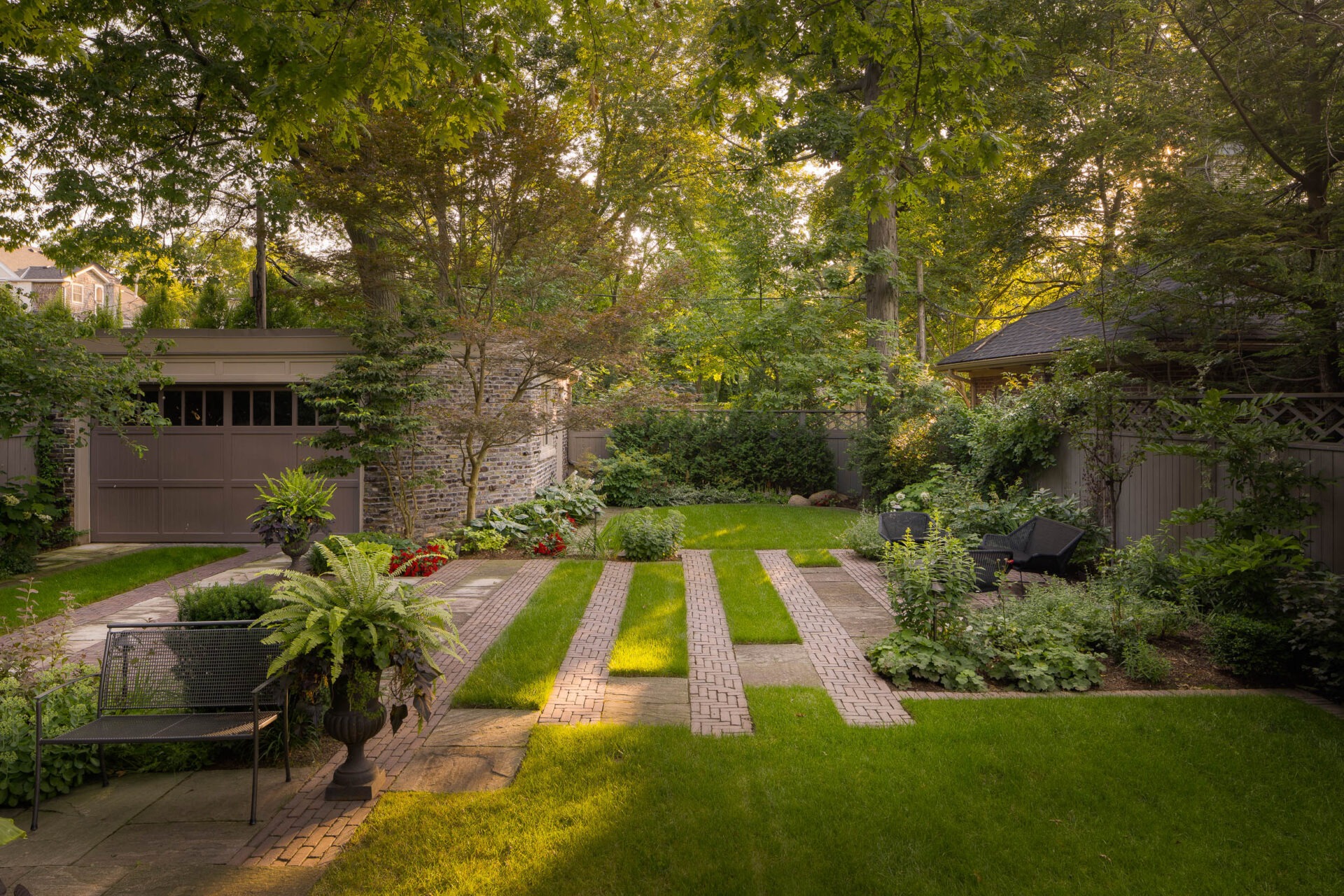 A tranquil garden with lush greenery features brick pathways, a bench, patio chairs, plants, and a garage under a canopy of trees.