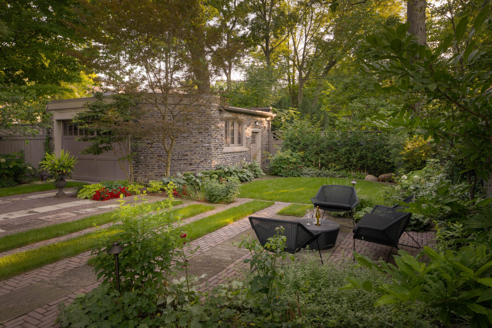 A serene garden scene with lush greenery, brick pathways, and a charming stone building surrounded by trees and outdoor seating. No persons visible.