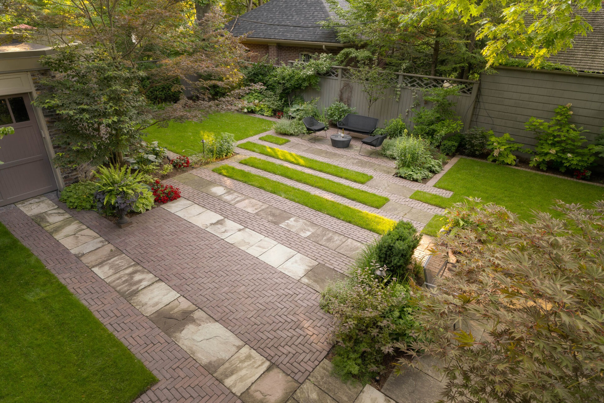 A well-maintained garden featuring brick pathways, lush greenery, and seating area with chairs and a table, surrounded by trees and plant beds.