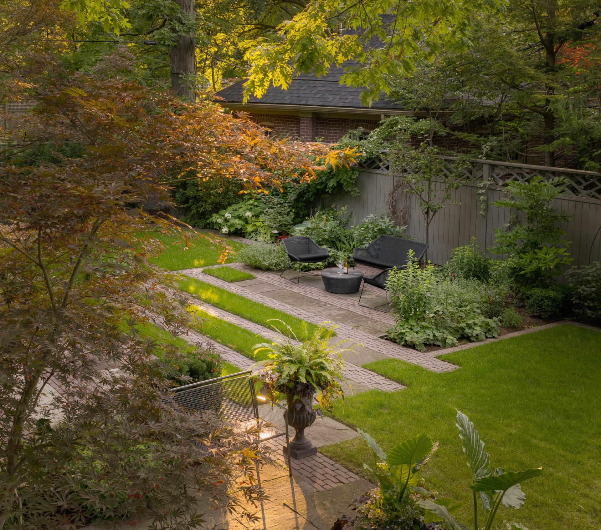 A peaceful, sunlit garden features lush greenery, paved paths, and two black chairs with a table, enclosed by a wooden fence.