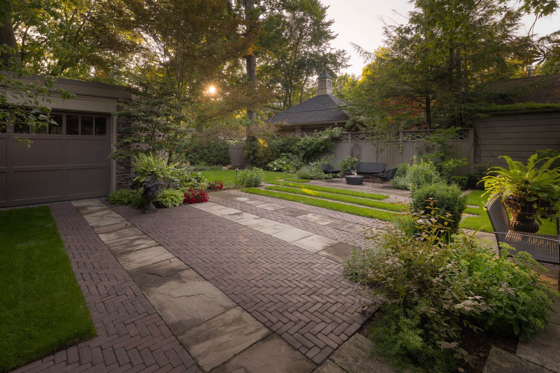 A serene garden with brick and stone pathways, lush greenery, and outdoor seating; sunlight filters through tall trees, creating a peaceful ambiance.