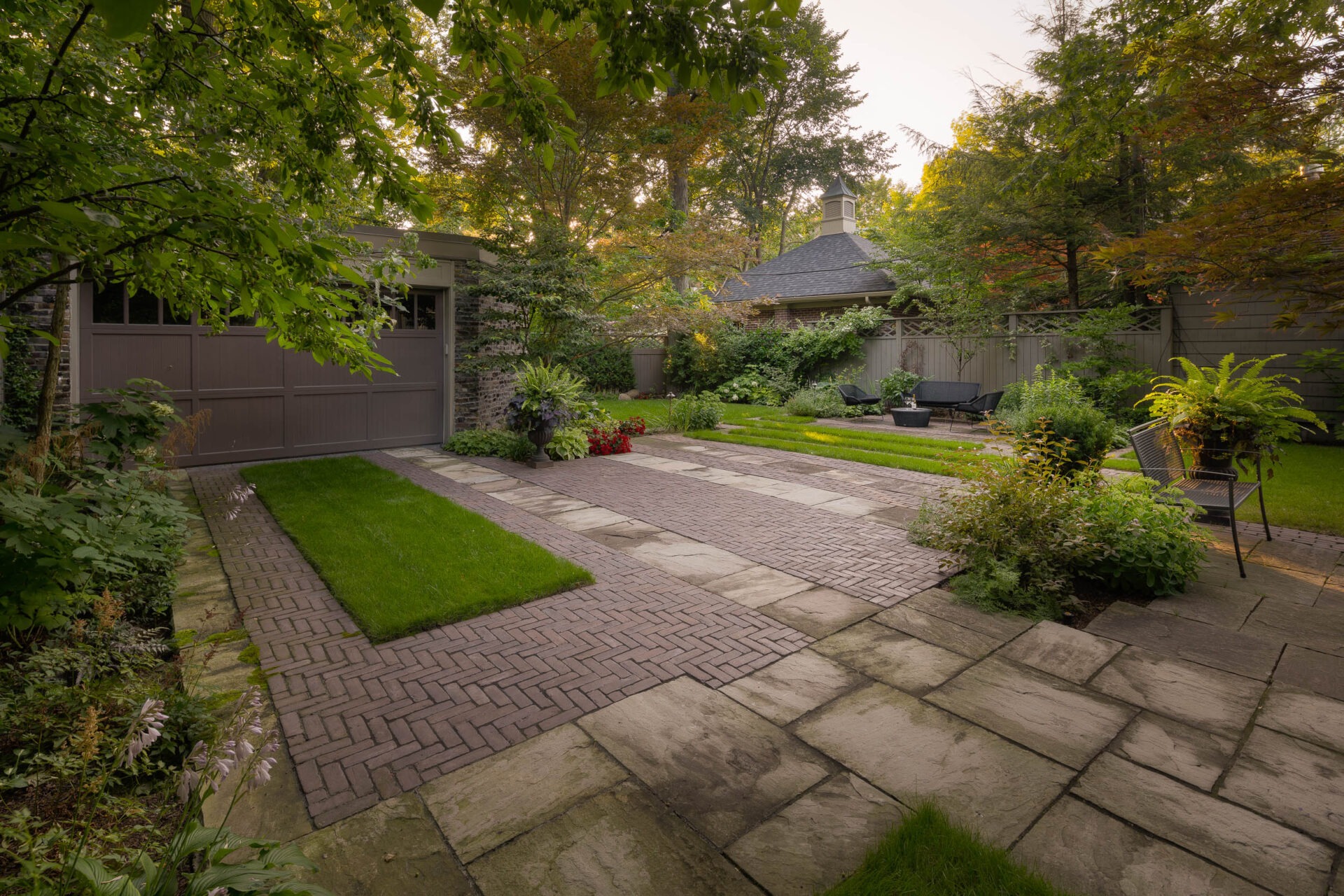 A serene backyard with brick and stone pathways, surrounded by lush greenery, a garage, and cozy seating area for relaxation.