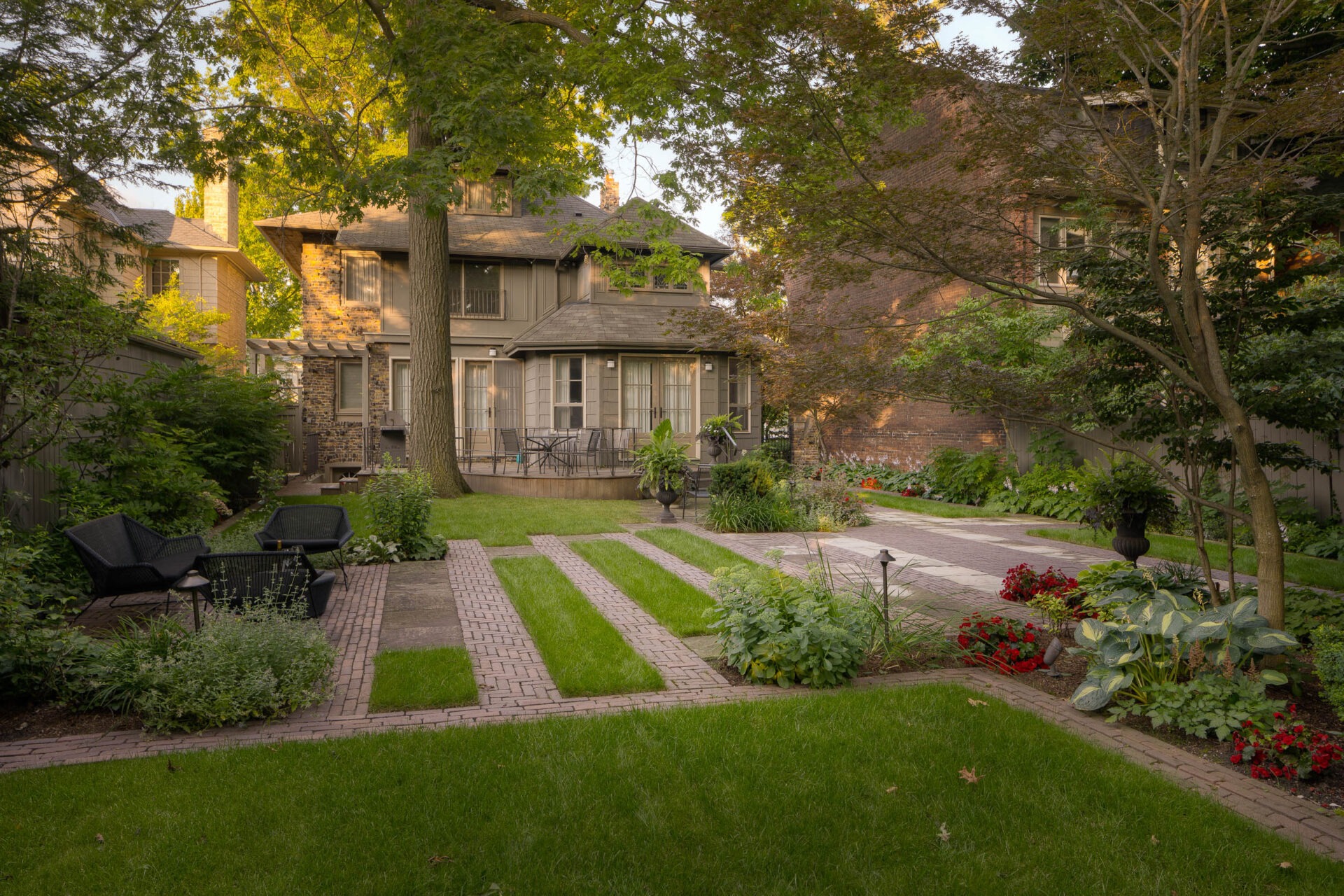 A lush garden with patio chairs and tables, surrounded by tidy green grass and plants, leads to a two-story house in the background.