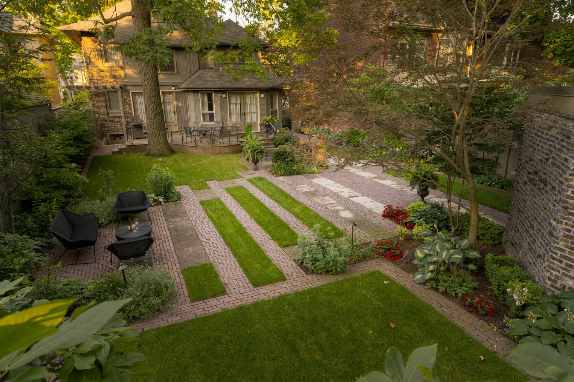 Lush garden with brick pathways, patio furniture, and greenery surrounding a traditional house. Trees provide shade, creating a serene, inviting atmosphere.