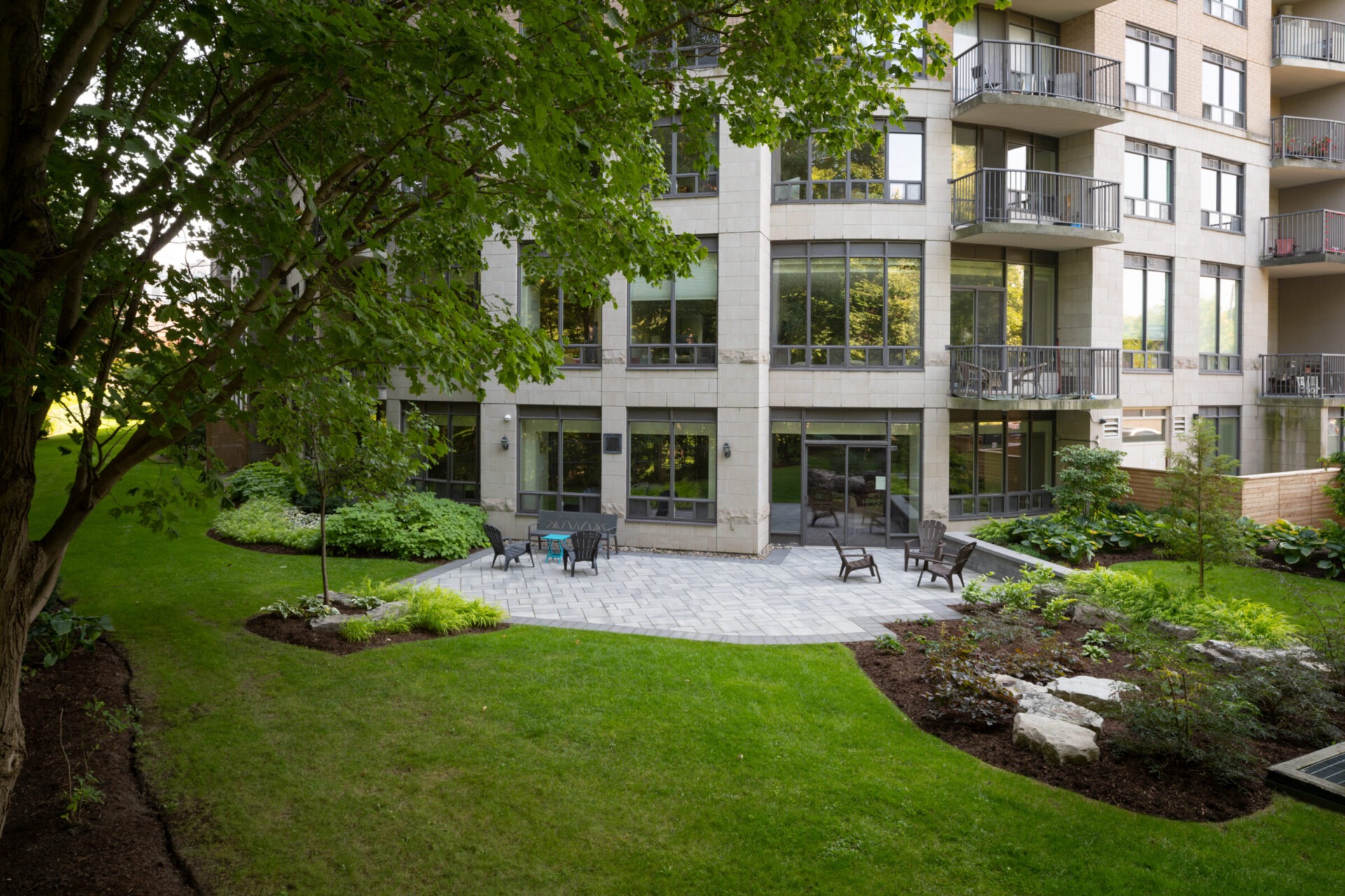 Modern apartment building with large windows and balconies, surrounded by trees and a landscaped garden with a stone patio and chairs.