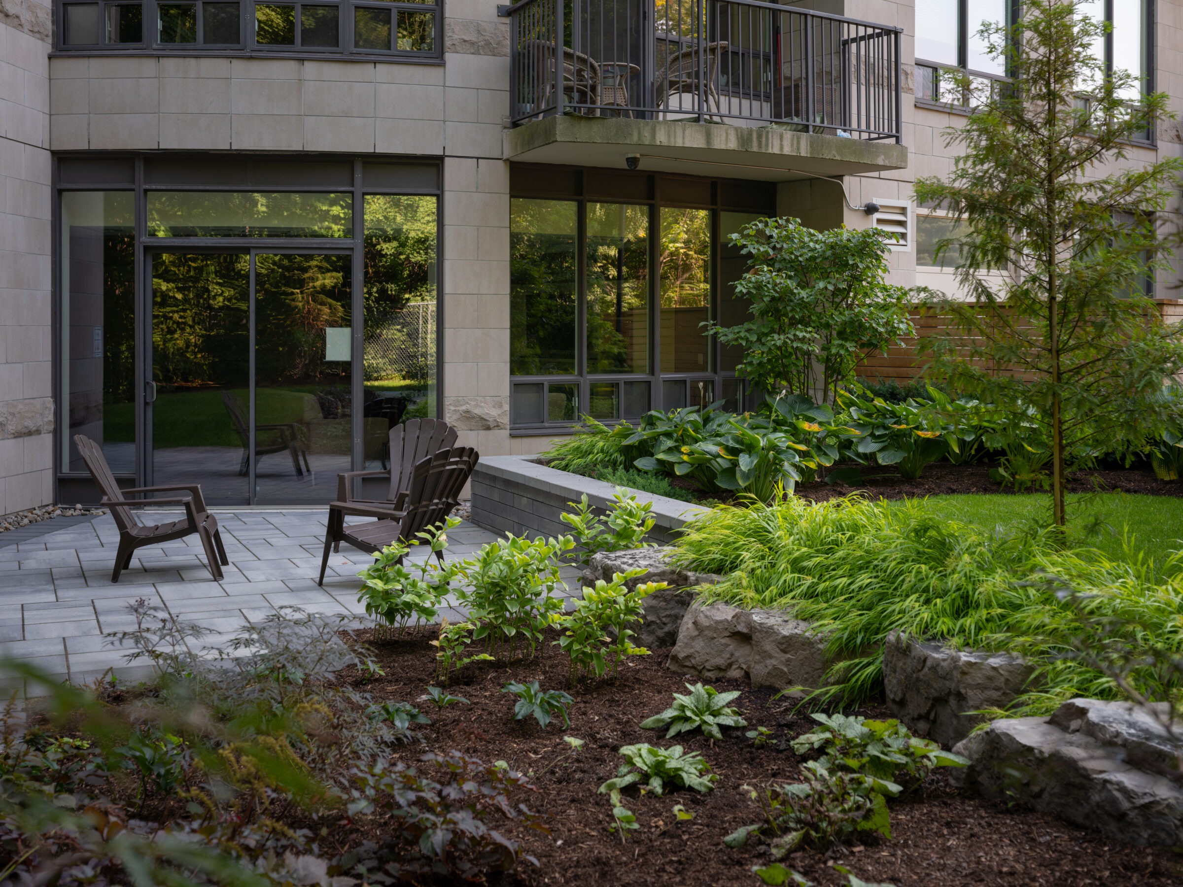 Modern patio with stone tiles, chairs, and lush greenery. Large windows reflect the light, creating a serene outdoor space with verdant plants.