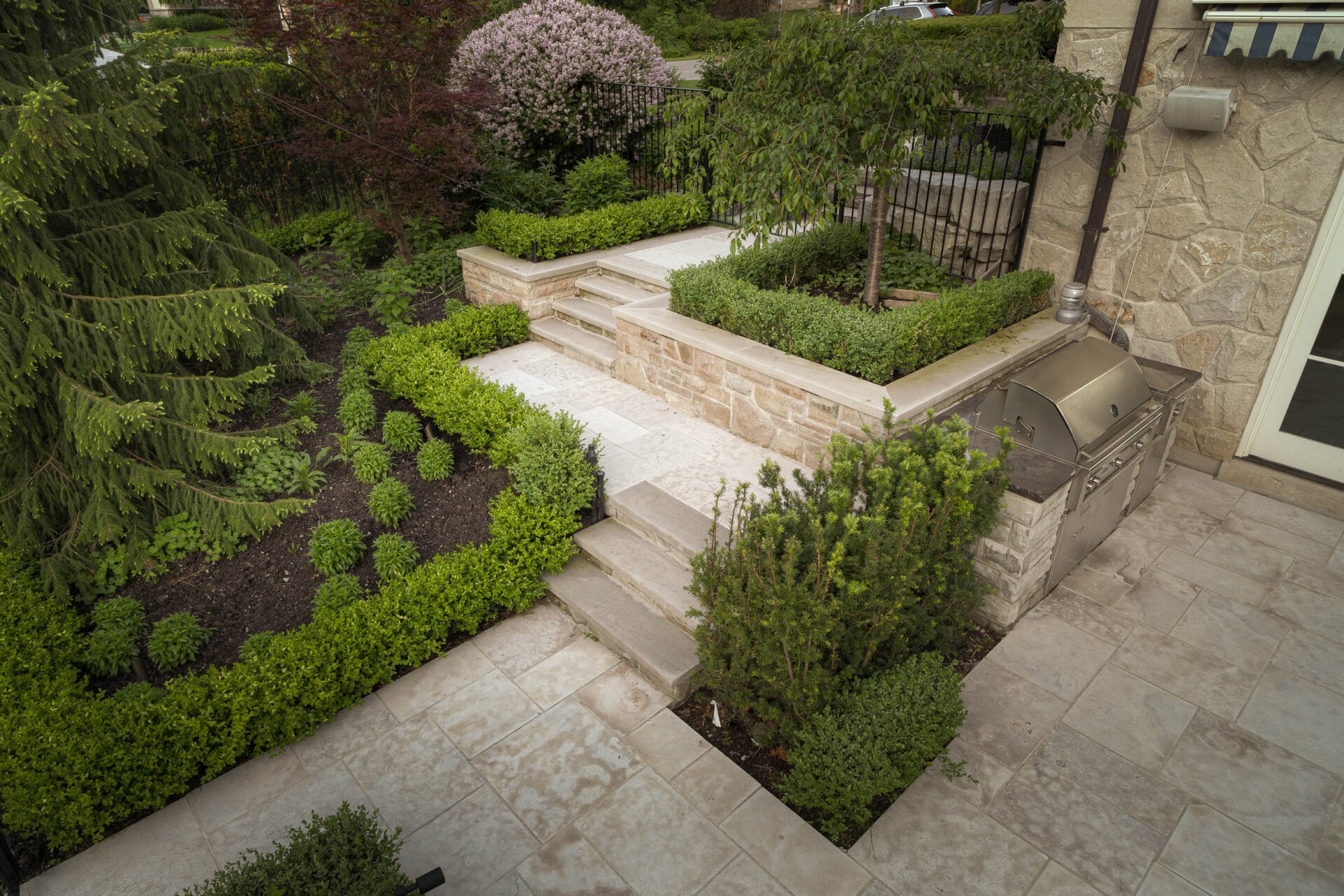 Elegant stone patio with stairs, lush greenery, and a barbecue grill. Well-maintained landscaping features various shrubs and a decorative stone wall.