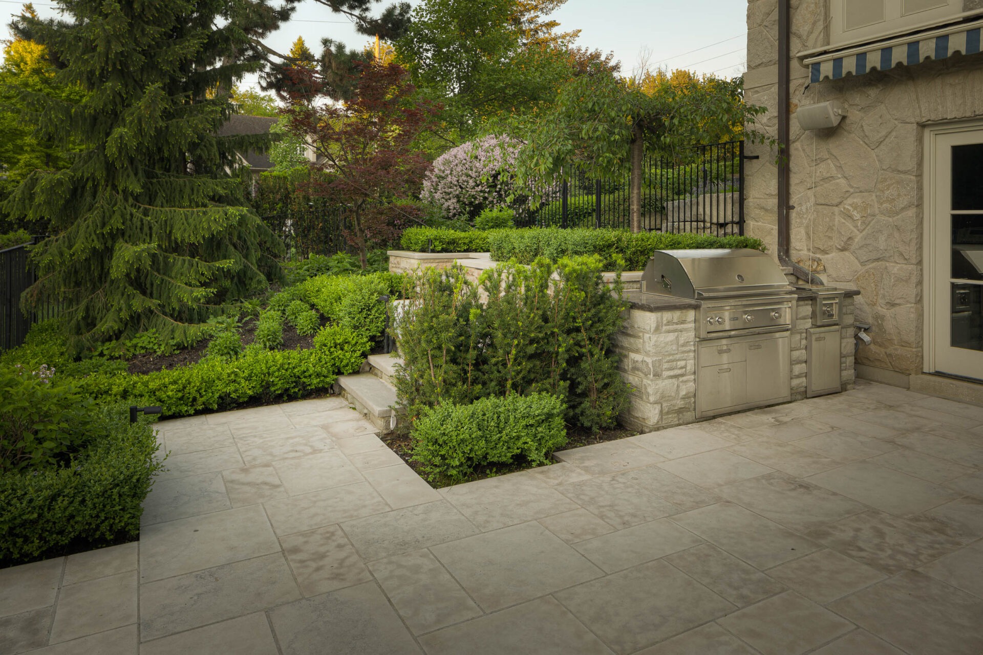 A stone patio with a built-in grill, surrounded by lush greenery and a variety of trees and shrubs.