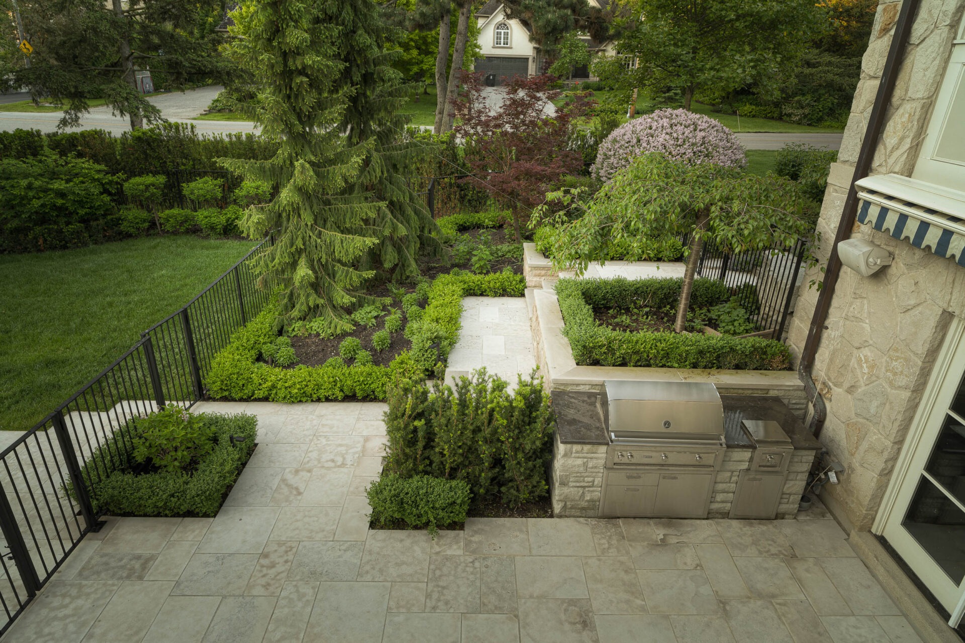 A landscaped backyard features a patio with a built-in grill, lush greenery, and a pathway, set beside a residential area.