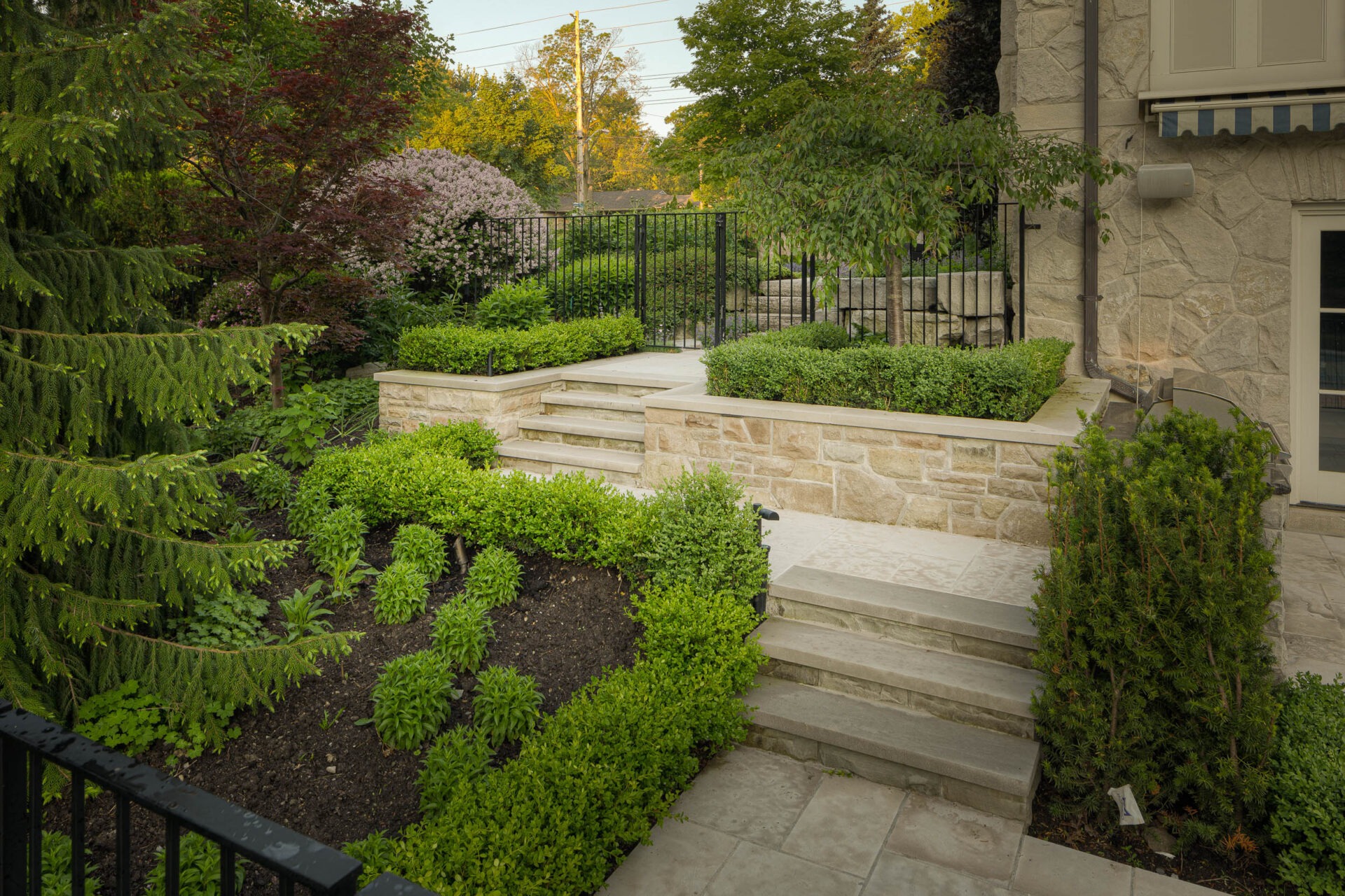 A beautifully landscaped garden features stone steps, lush greenery, and a stone wall, bordered by black fencing and vibrant foliage.