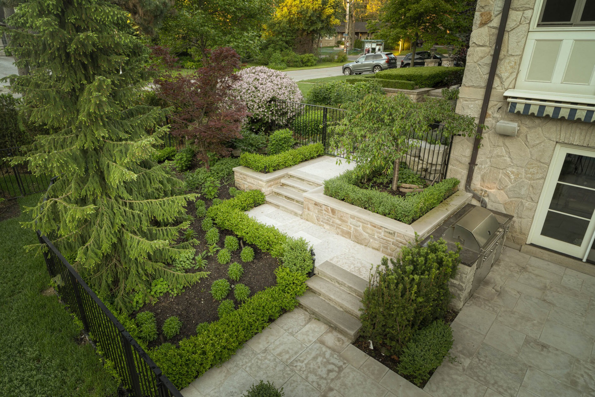 A well-manicured garden with stone steps, lush greenery, and a barbecue grill. A quiet residential street is in the background.