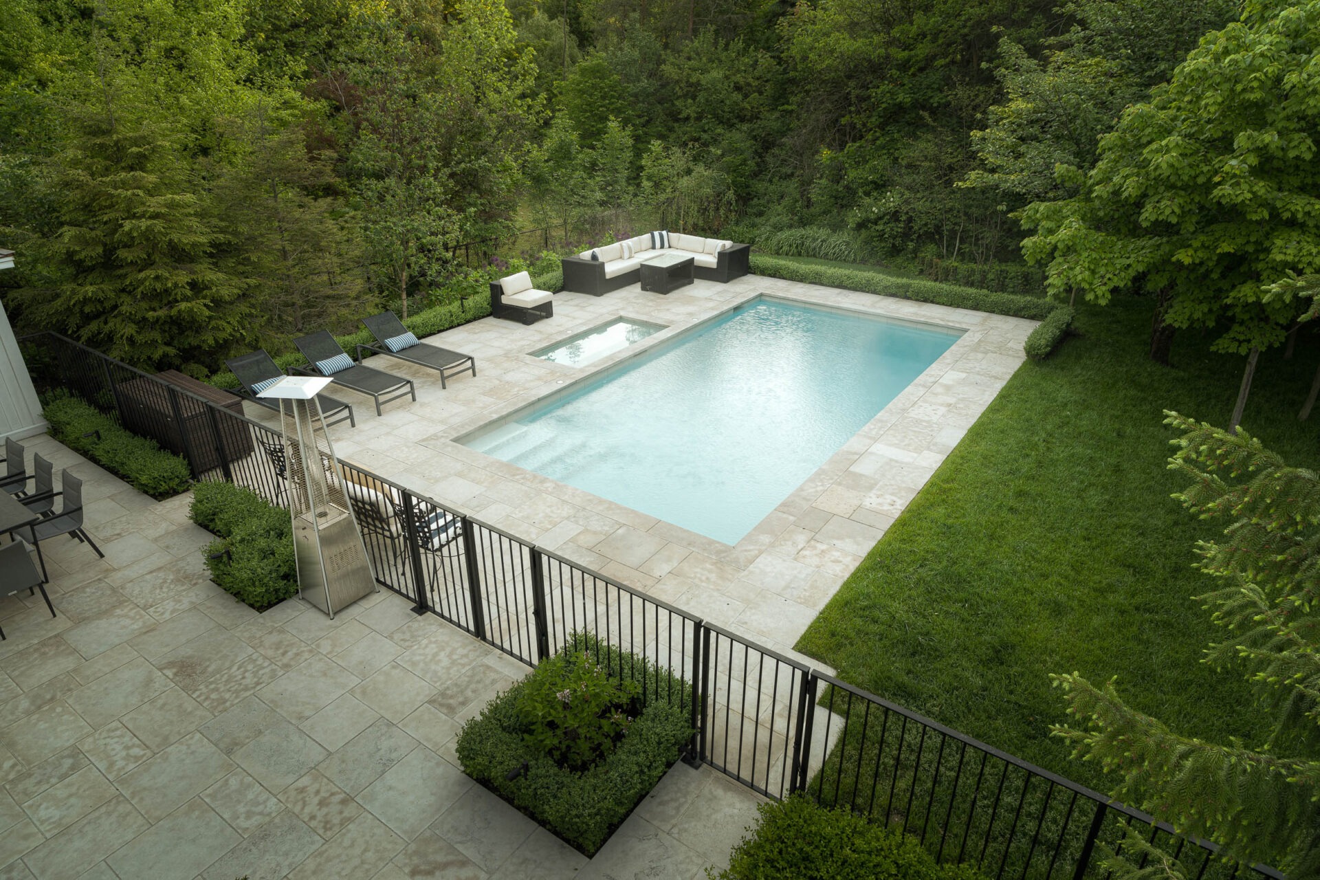 Aerial view of a serene backyard with a rectangular pool, surrounded by lush greenery, patio furniture, and trees.