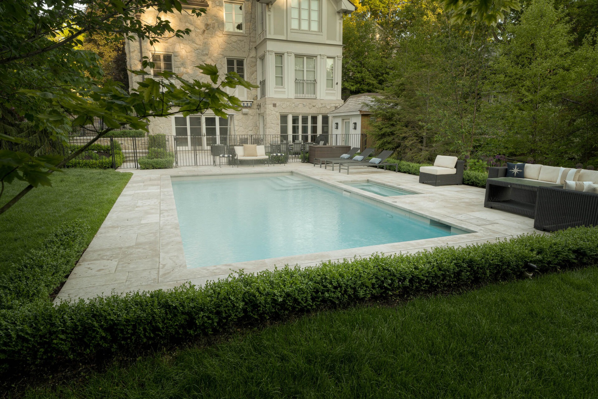 A luxurious backyard features a sparkling pool, surrounded by green landscaping and elegant seating, adjacent to a large stone house.