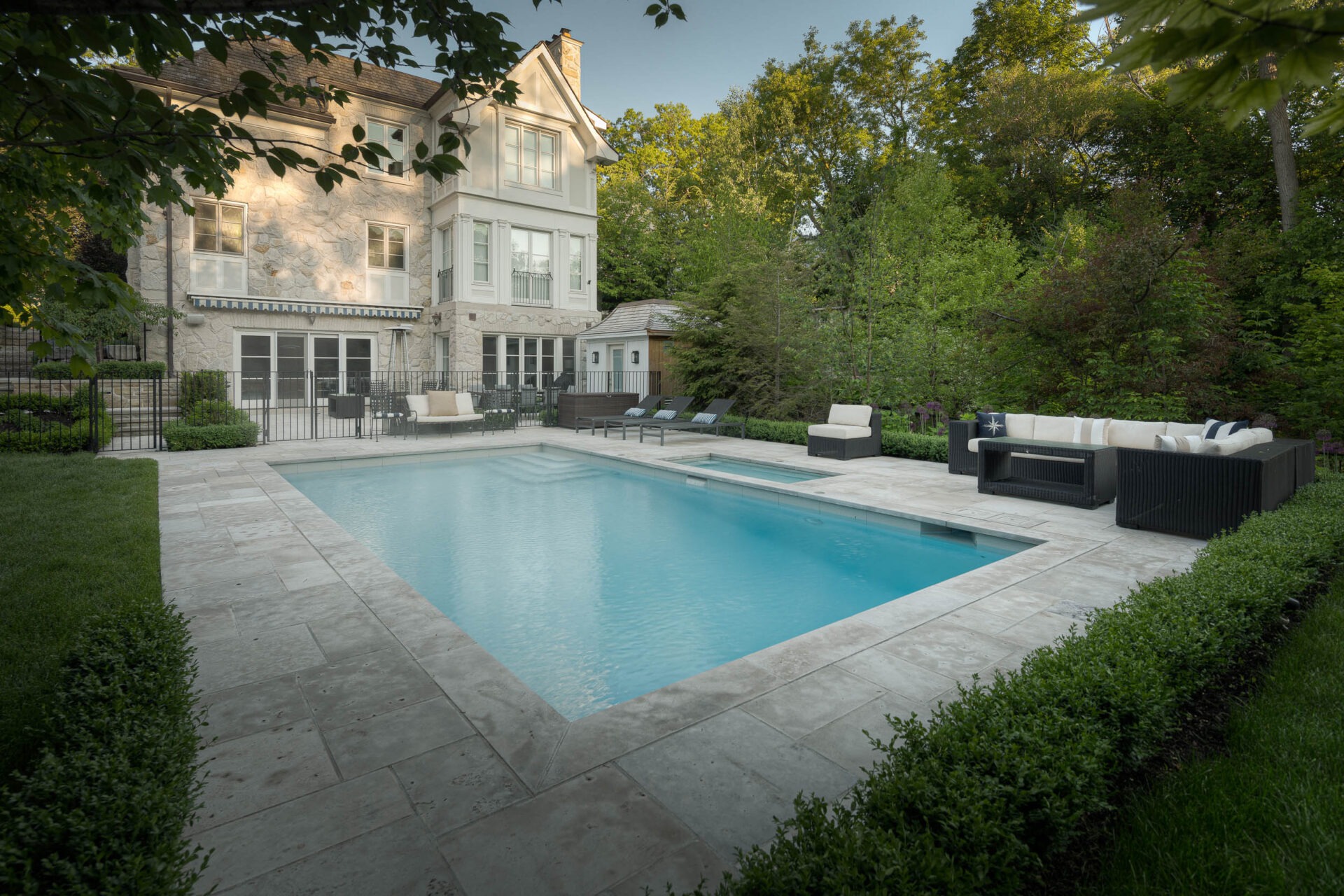 A stone house with large windows overlooks a rectangular pool surrounded by patio furniture and lush greenery in a tranquil setting.