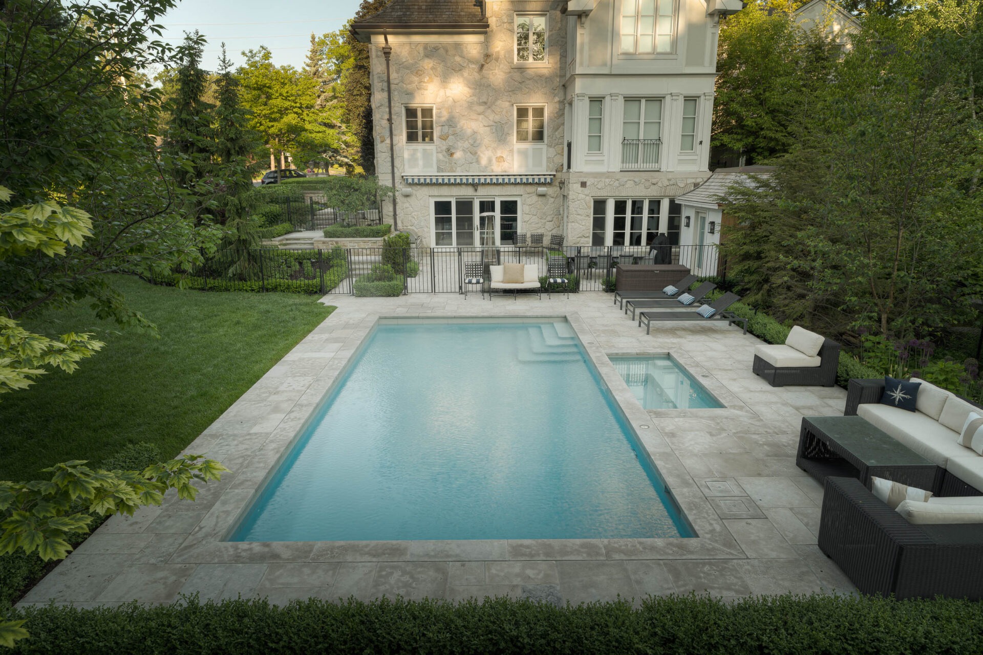 Elegant backyard with a swimming pool, hot tub, and stylish patio furniture set beside a large stone house, surrounded by lush greenery.