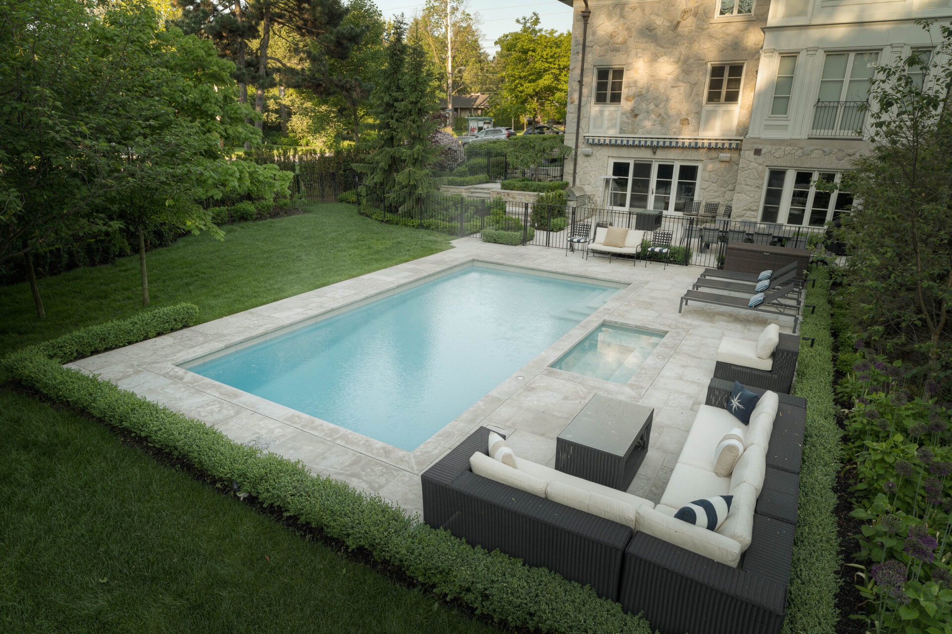 A serene backyard with a rectangular pool, surrounded by lush greenery, adjacent to a stone house. Patio furniture is neatly arranged.