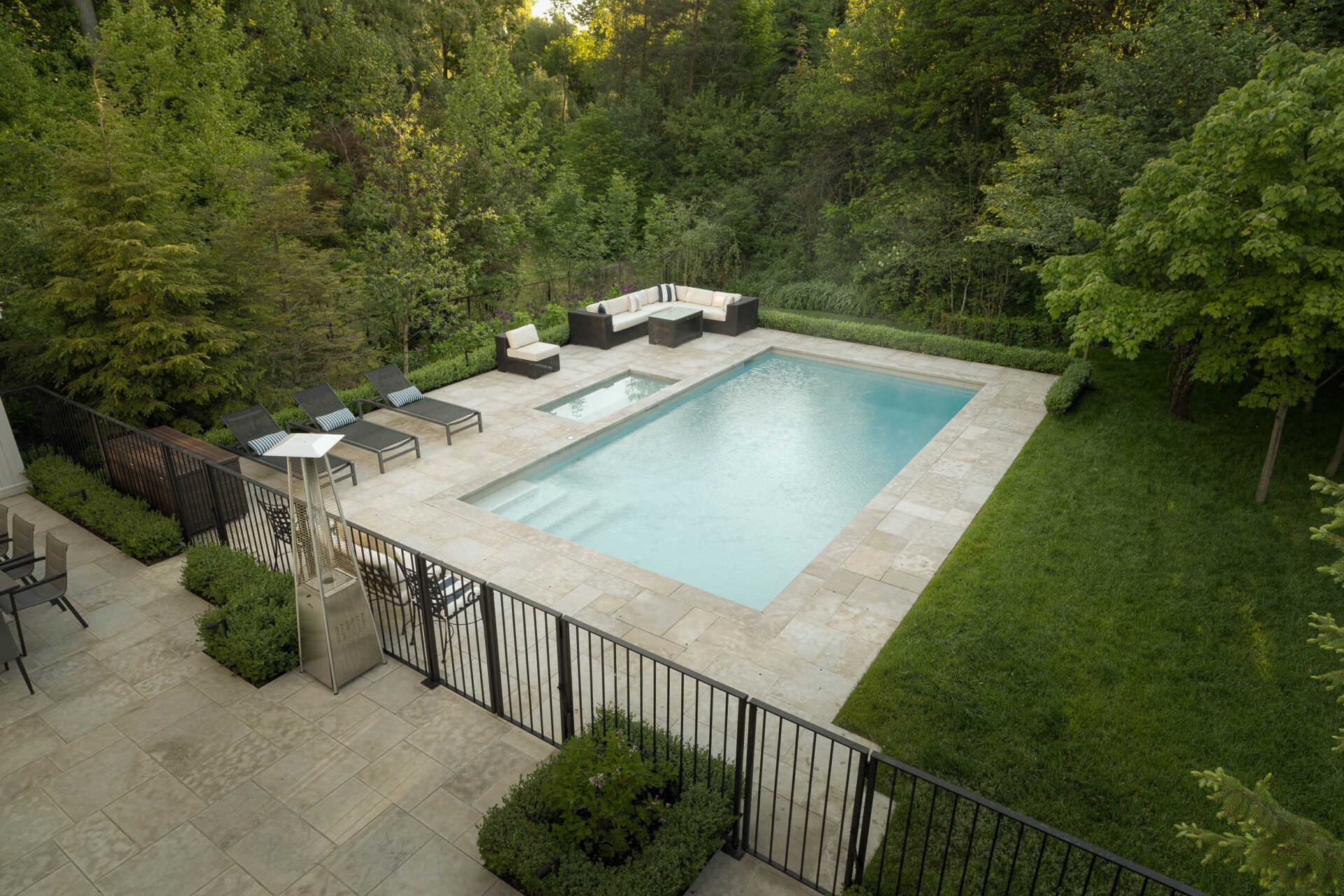 A serene backyard features a rectangular swimming pool surrounded by greenery, lounge chairs, and an outdoor seating area, enclosed by black fencing.